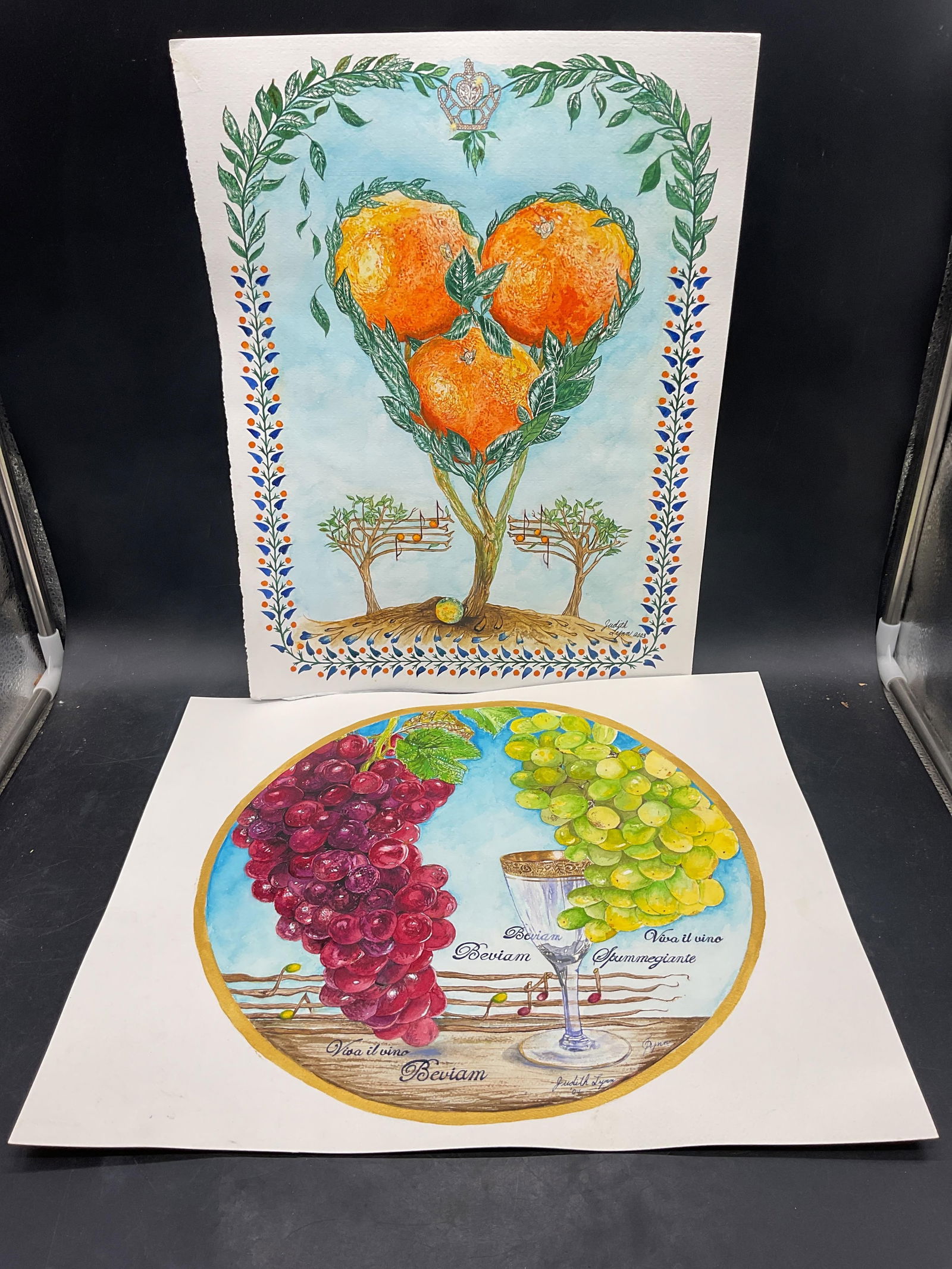 Lot 2 Judith Lynn Fruit Music Watercolor Paintings: Lot of 2 watercolor paintings on paper. Signed Judith Lynn. Orange tree and music notes, grapes, wine glass and music notes. Orange painting measures approx 12 x 16 inches. Some wear to each consisten