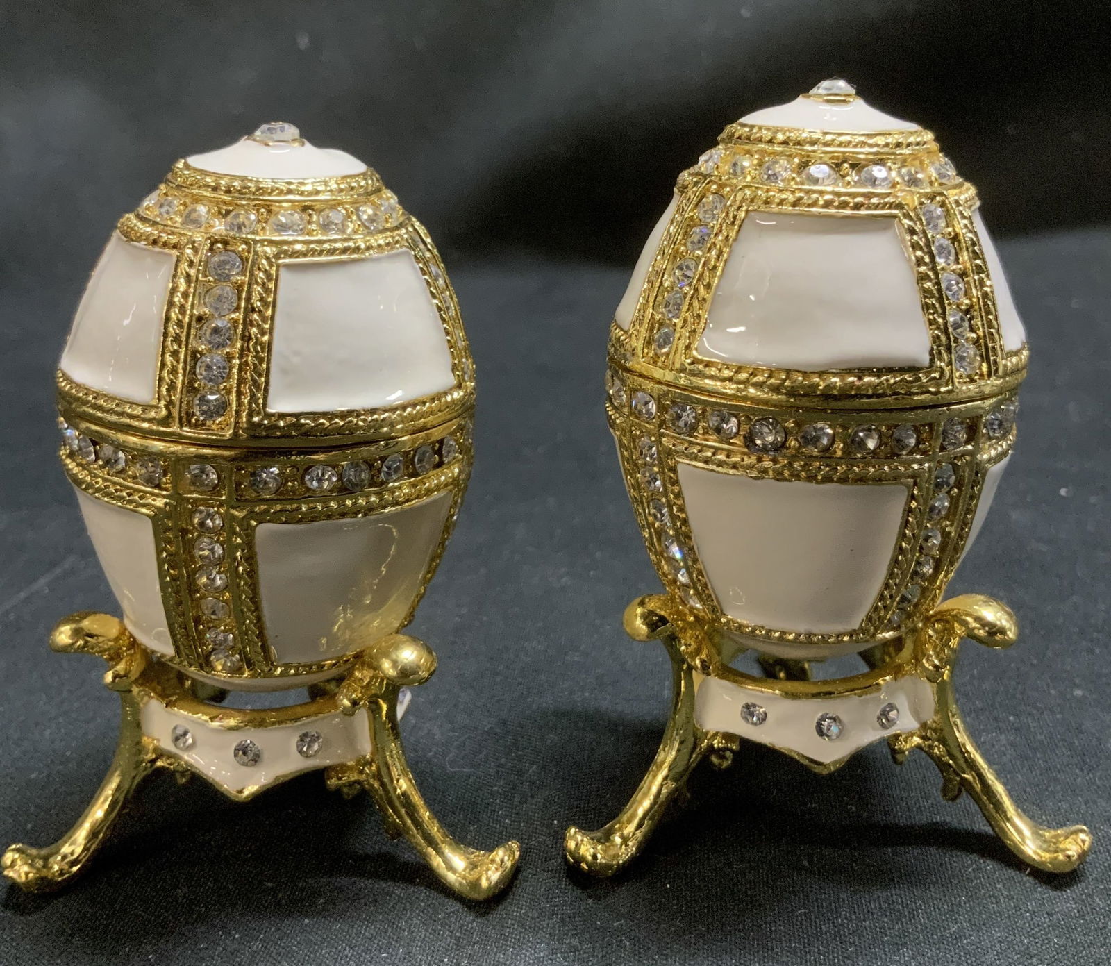 Pair Enameled & Rhinestone Trinket Bx W Stand: Lot includes, 2 enameled & rhinestone egg trinket boxes. Gold toned. Comes with matching stand. Surface wear consistent to age, use & exposure. Measurements: length 1.5in x width 1.5in x height