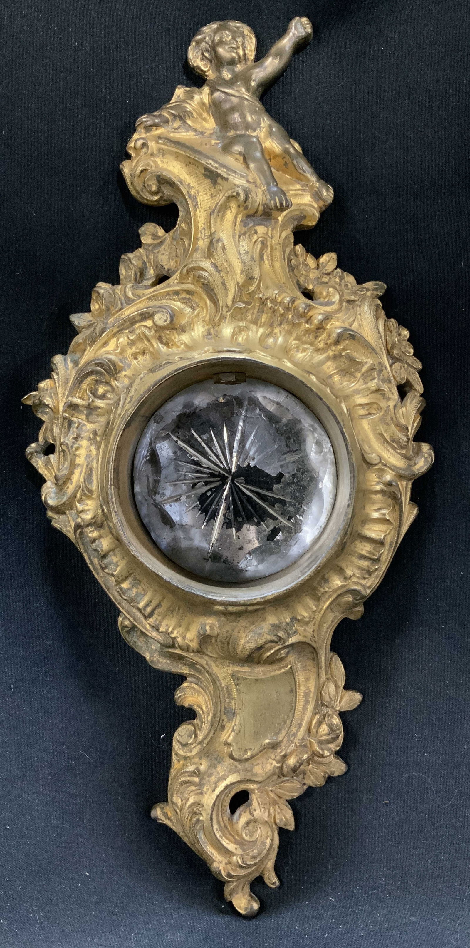 Rocco Revival Style Gilt Metal Cherub Wall Decor: Vintage ornate rococo revival style wall decor featuring a cherub seated atop & floral motifs. Glass insert in center. Gilt accents. Sold as is. Signed & marked on underside. Visible numbers. Surface