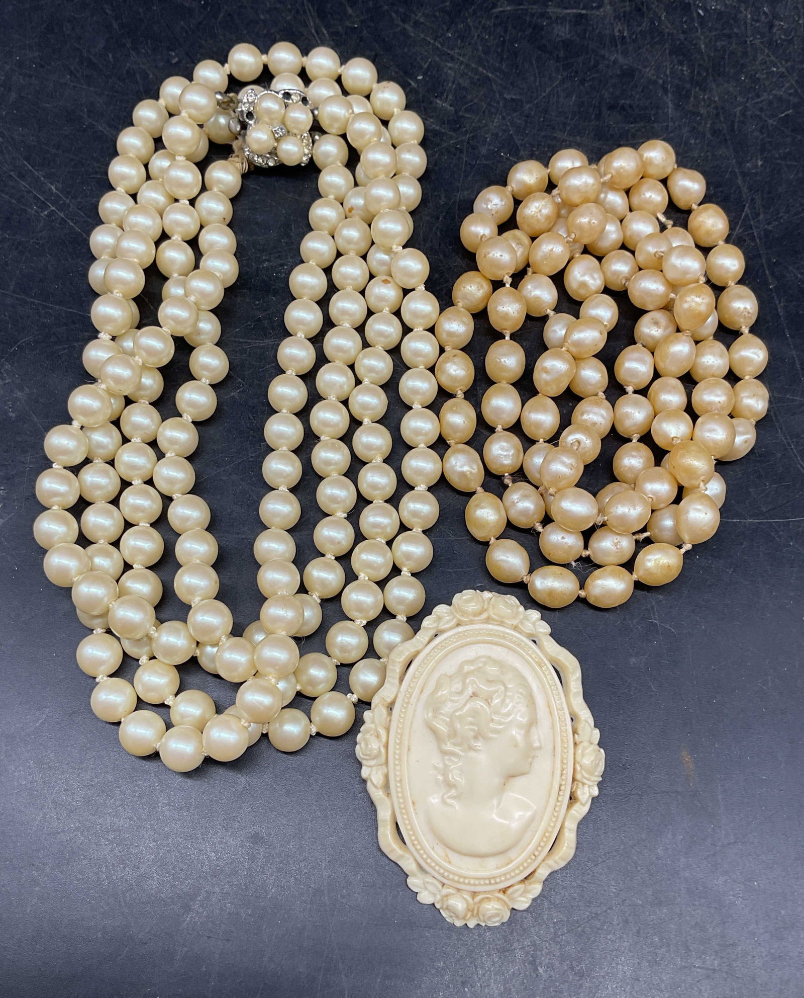 Lot 3 Faux Pearl Necklaces, Cameo Brooch: Lot of 3 jewelry pieces. 2 faux pearl beaded necklace. Longer drop length approx 17 inches. White acrylic or resin cameo brooch. Measures approx. 2.25 x 1.75 inches. Some scratching. Crystals on clasp