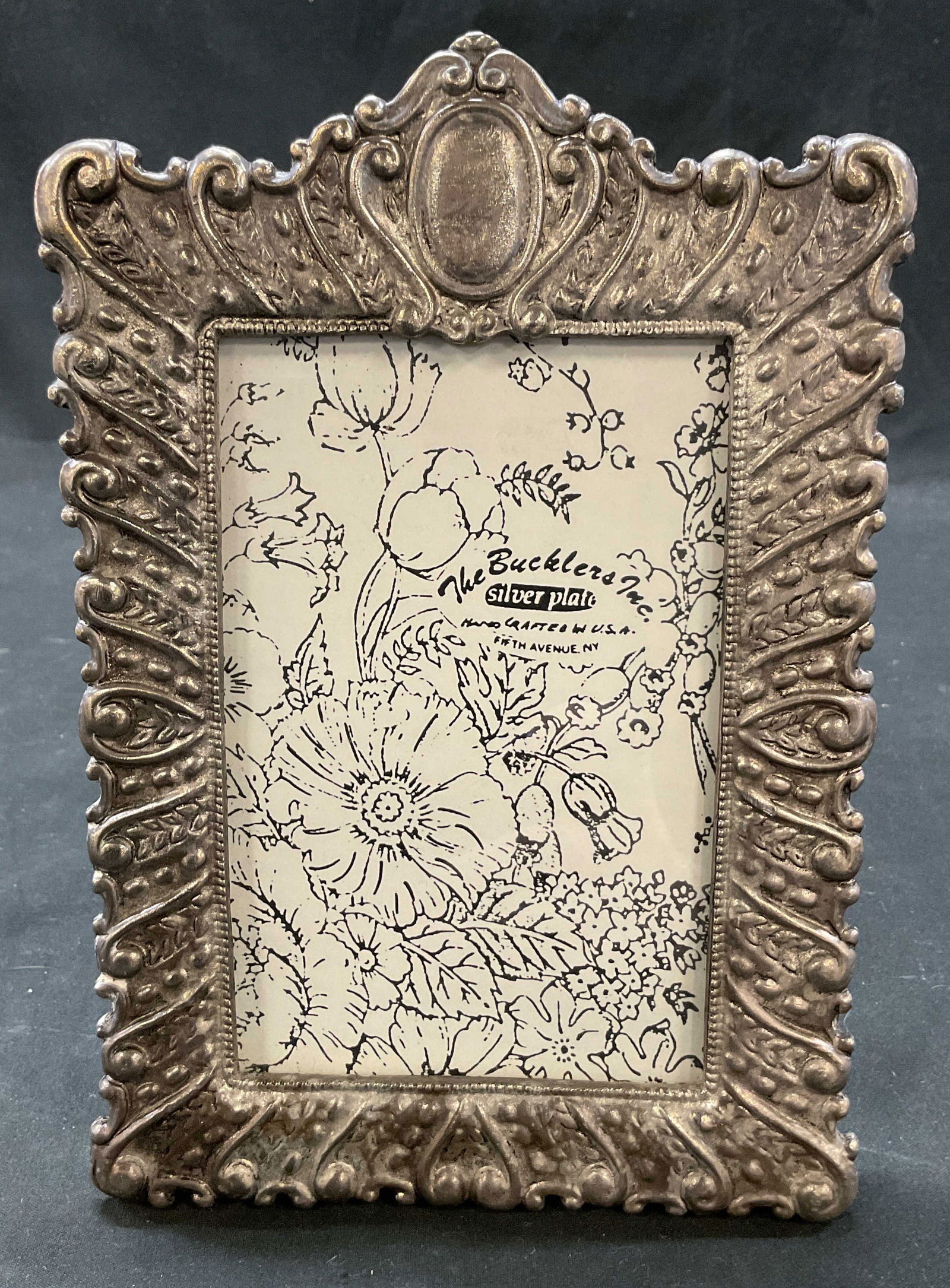 Bucklers Inc Silver Plated Picture Frame USA: The Bucklers Inc silver plated picture frame. Reads The Bucklers Inc Silver Plate Hand Crafted In USA Fifth Avenue NY. Embossed scroll motif pattern. Rectangular shape. Measures approx 5.5 x 7 inches.