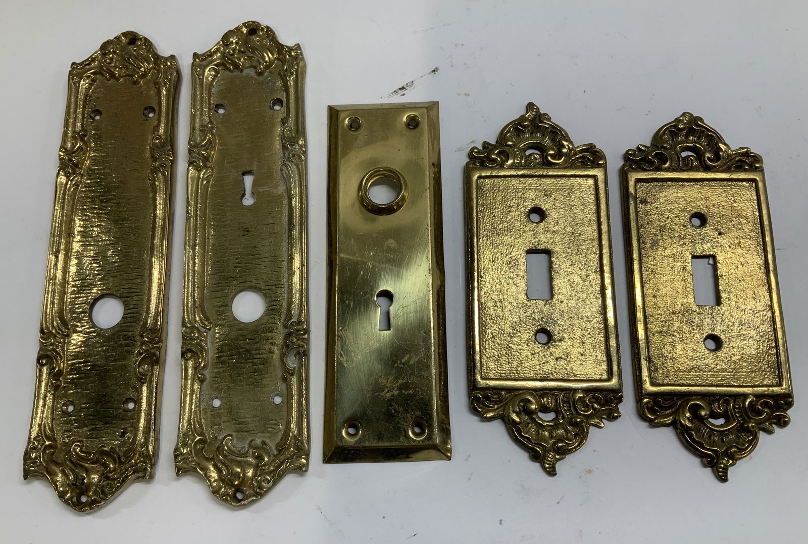 Lot 5 Brass Door Escutcheon Plates & Covers: Lot includes, 3 brass door escutcheon plates & 2 light switch covers. Features ornate detailing. Victorian style. Surface wear consistent to age, use & exposure. Measurements: length 9.5in x width 2.3