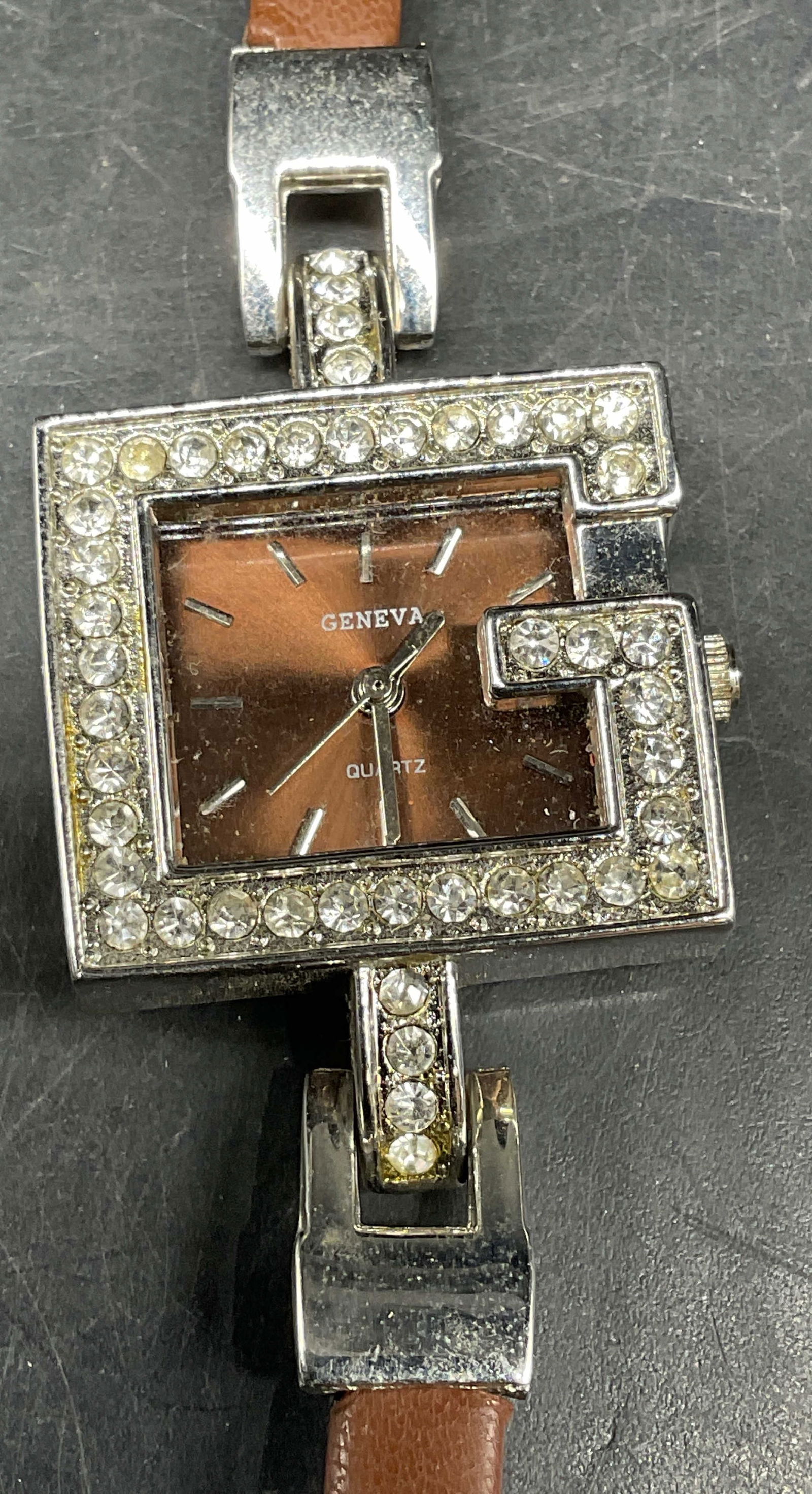 Geneva Silver Tone Crystal G Watch: Geneva silver tone and crystal G motif watch with brown faux leather strap. Measures approx. 8 inches long. Strap needs replacement. Not tested. Some scratching. Jewelry, estate jewelry, costume jewel