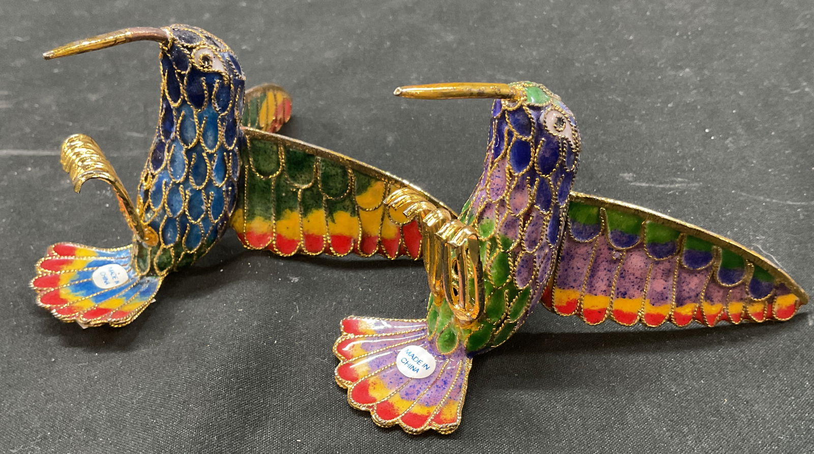 Pair Chinese Cloisonne Hummingbird Figures: Lot of 2 Chinese cloisonne hummingbird figures. Hummingbird figure, enameled metal. Long wings and small body with feet. Long beak. Gold toned and varying colors. Measures approx 3 x 4 x 2.5 inches. S