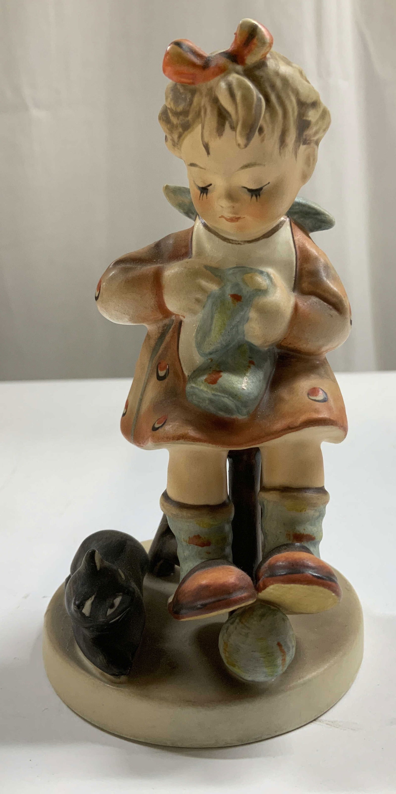 Vtg Goebel Mothers Helper Figurine W. GERMANY: Vintage Goebel Mothers Helper figurine depicts a young girl sitting on a chair. Small cat resting beside her. Ceramic material. Signed & marked on underside. W. GERMANY. Surface wear consistent to age