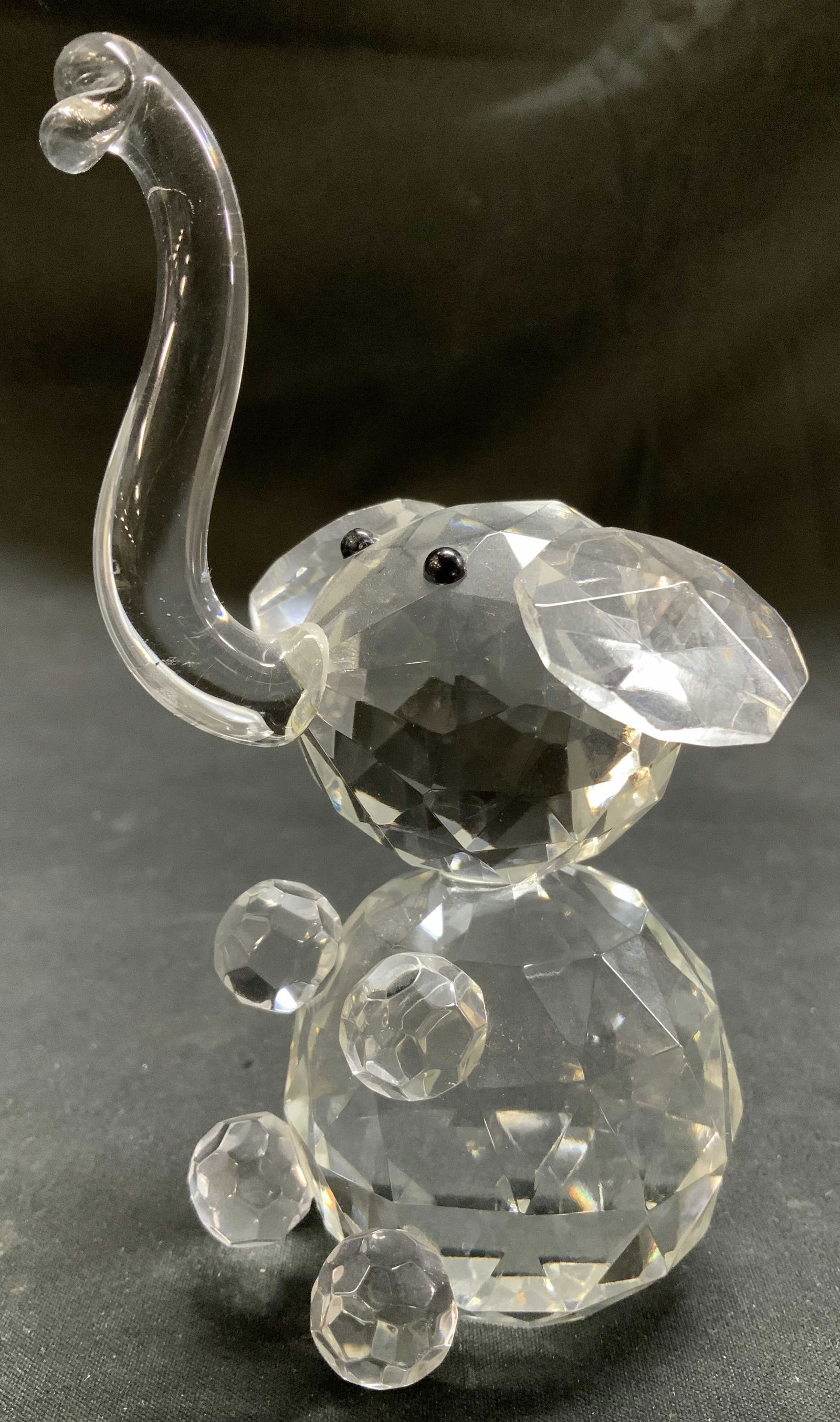 Clear Crystal Elephant Figurine: Crystal elephant figurine in a sitting pose with its trunk upwards. Features black eyes. Surface wear consistent to age, use & exposure. Measurements: length 2.7in x width 2.5in x height 4.5in. Clear
