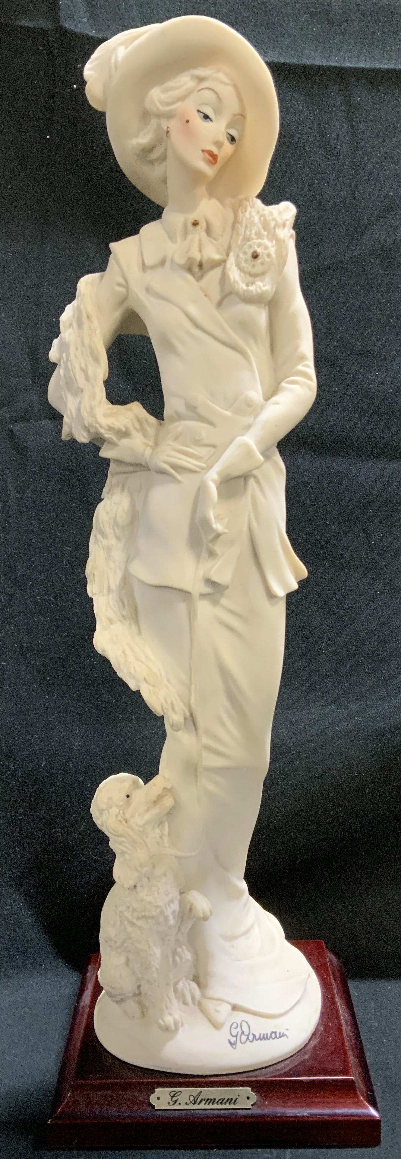 1987 Sgnd Giuseppe Armani Lady W Poodle Fig ITALY: 1987 Giuseppe Armani figurine depicting an elegant woman wearing a large hat. Small poodle by her feet. Figurine is mounted on a dark wooden base with a gold toned plaque. Reads, G. Armani. Signed & m