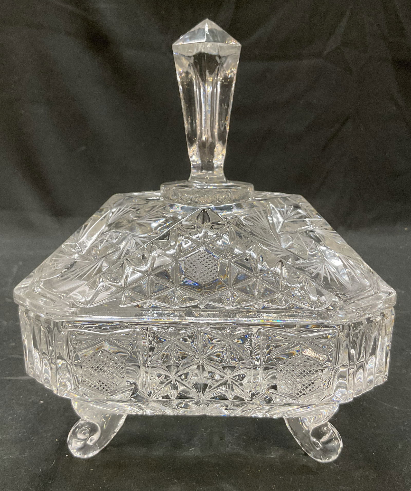 Vintage Footed Cut Crystal Lidded Candy Dish: Vintage footed cut crystal lidded candy dish. Four small scroll feet on underside. Square shape with faceted corners. Cut designs featuring geometric patterns, starbursts, fan patterns and criss cross