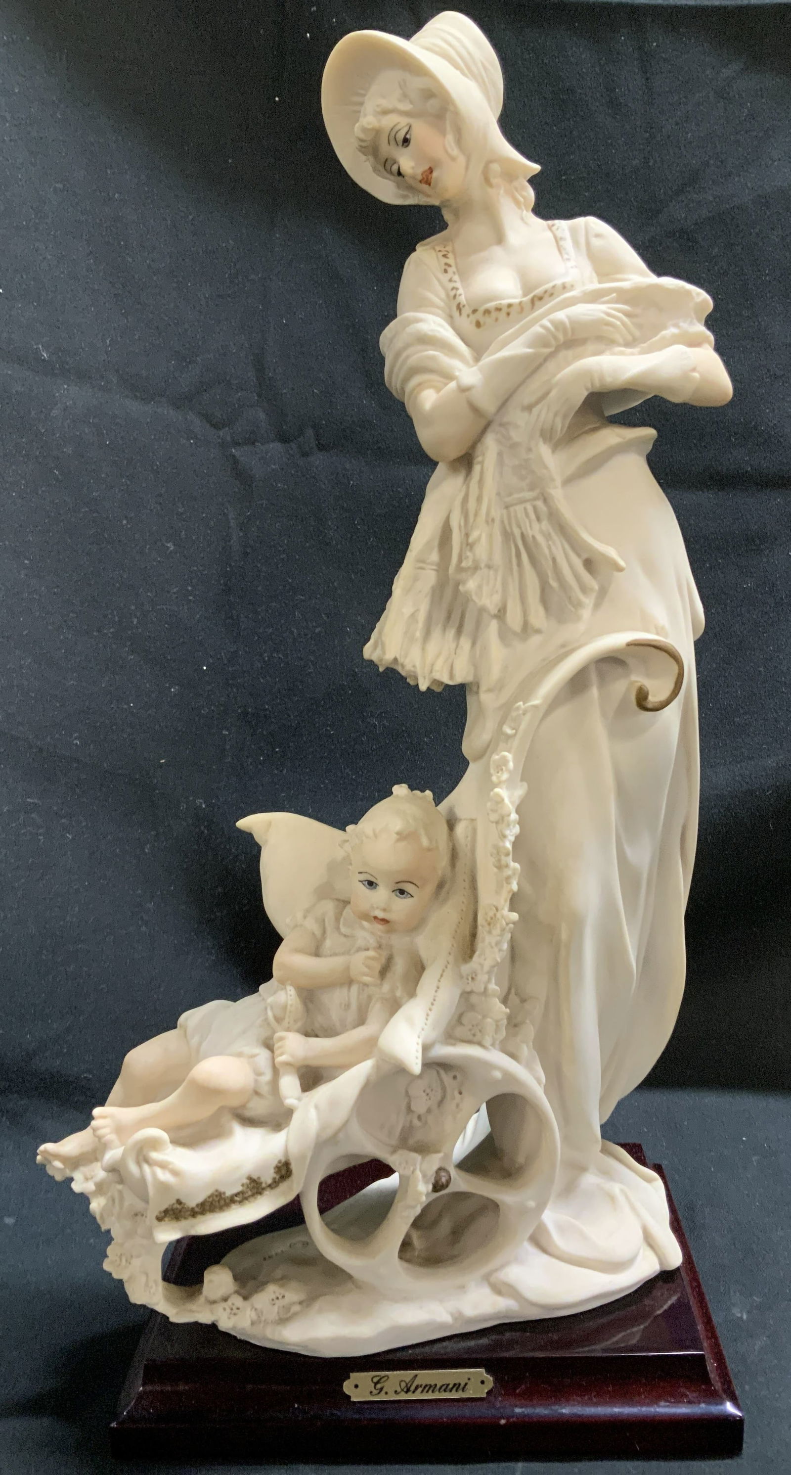 1987 Sgnd Giuseppe Armani Maternity Fig ITALY: 1987 Giuseppe Armani figurine depicting an elegant woman wearing a hat & pushing a baby carriage. Intricate detailing. Gilt accents. Figurine is mounted on a dark wooden base with a gold toned plaque.