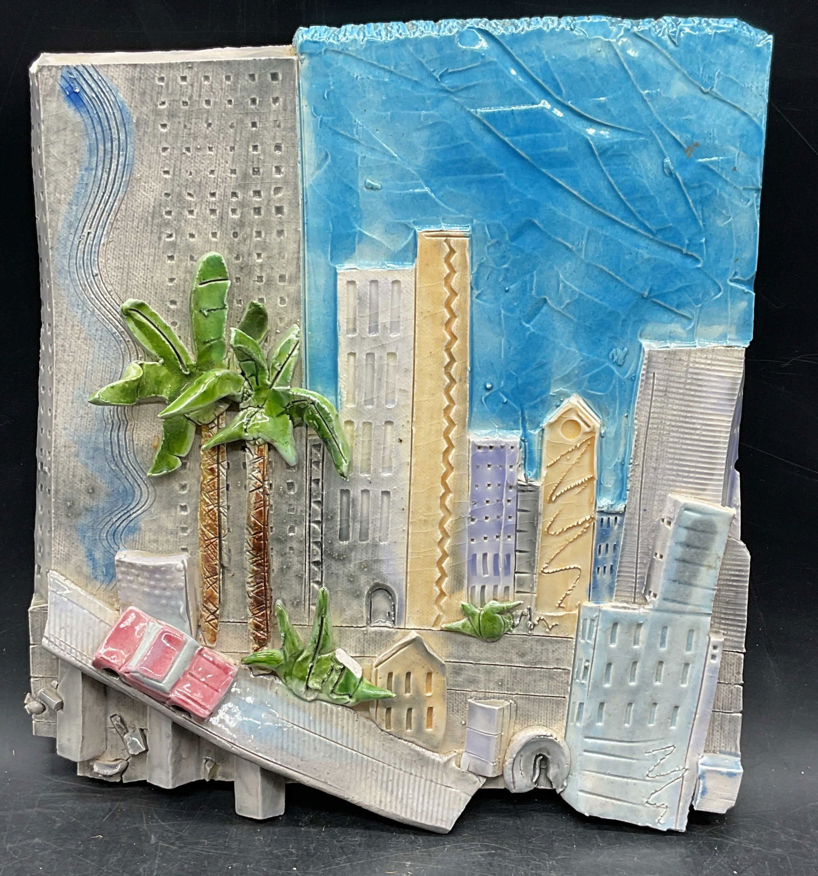 Signed Spiff 3D Urban Cityscape Will Plaque: Signed 3D relief urban cityscape wall plaque. Palm trees, car, and buildings. Signed Spiff, dated 1988. Measures approx. 9 x 8.5 inches. Piece has weight. Some scratching and small chips. Wall plaque,