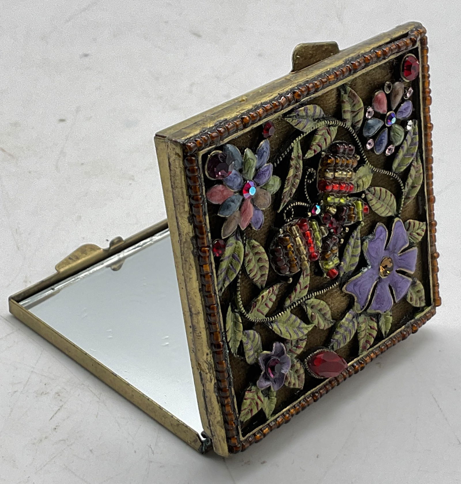 Vintage Enameled Metal Mirror Compact: Vintage enameled metal mirror compact. Gold toned. Square shape. Enameled with butterfly, floral, and leaf motifs. Beaded accents. Varying colors. Beaded border. Measures approx 2.5 x 2.5 inches. Surf