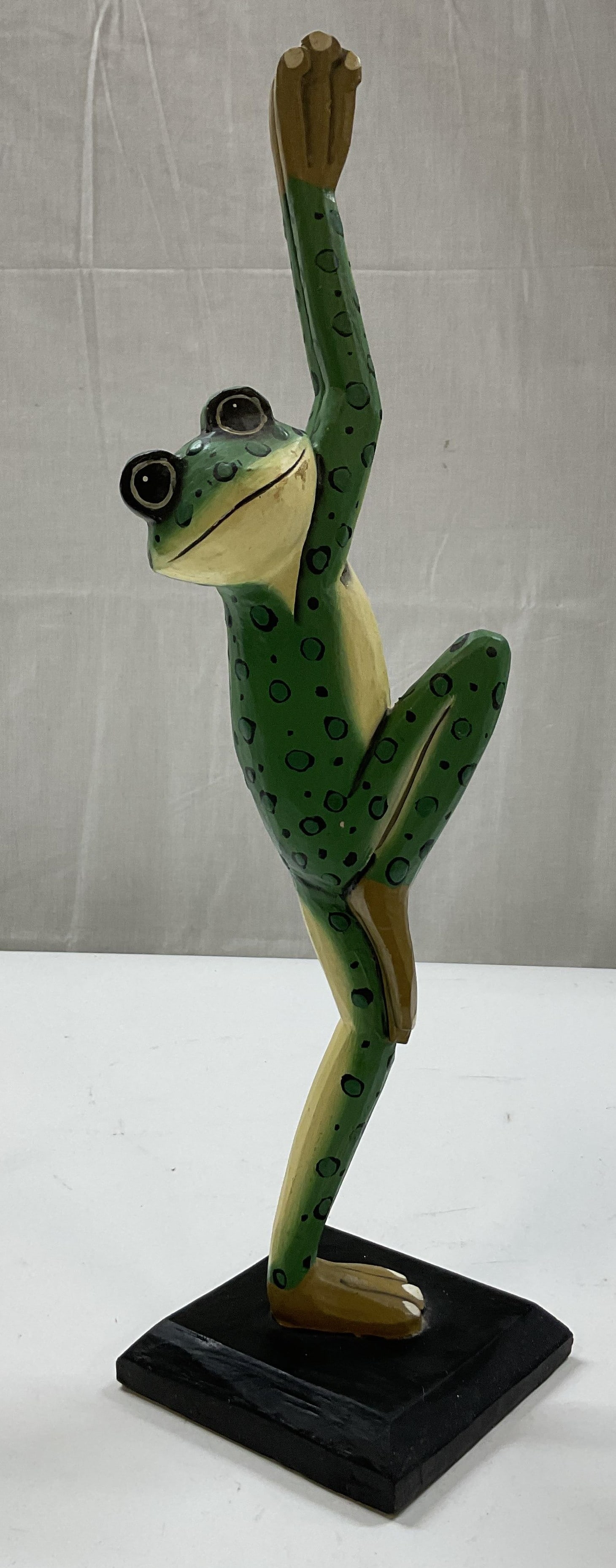 Hand Carved Wooden Frog Figure: Hand carved and hand painted wooden frog figure. Square black base. Frog figure standing on one foot with arms and other leg raised. Hand painted features in green and black. Measures approx 4.5