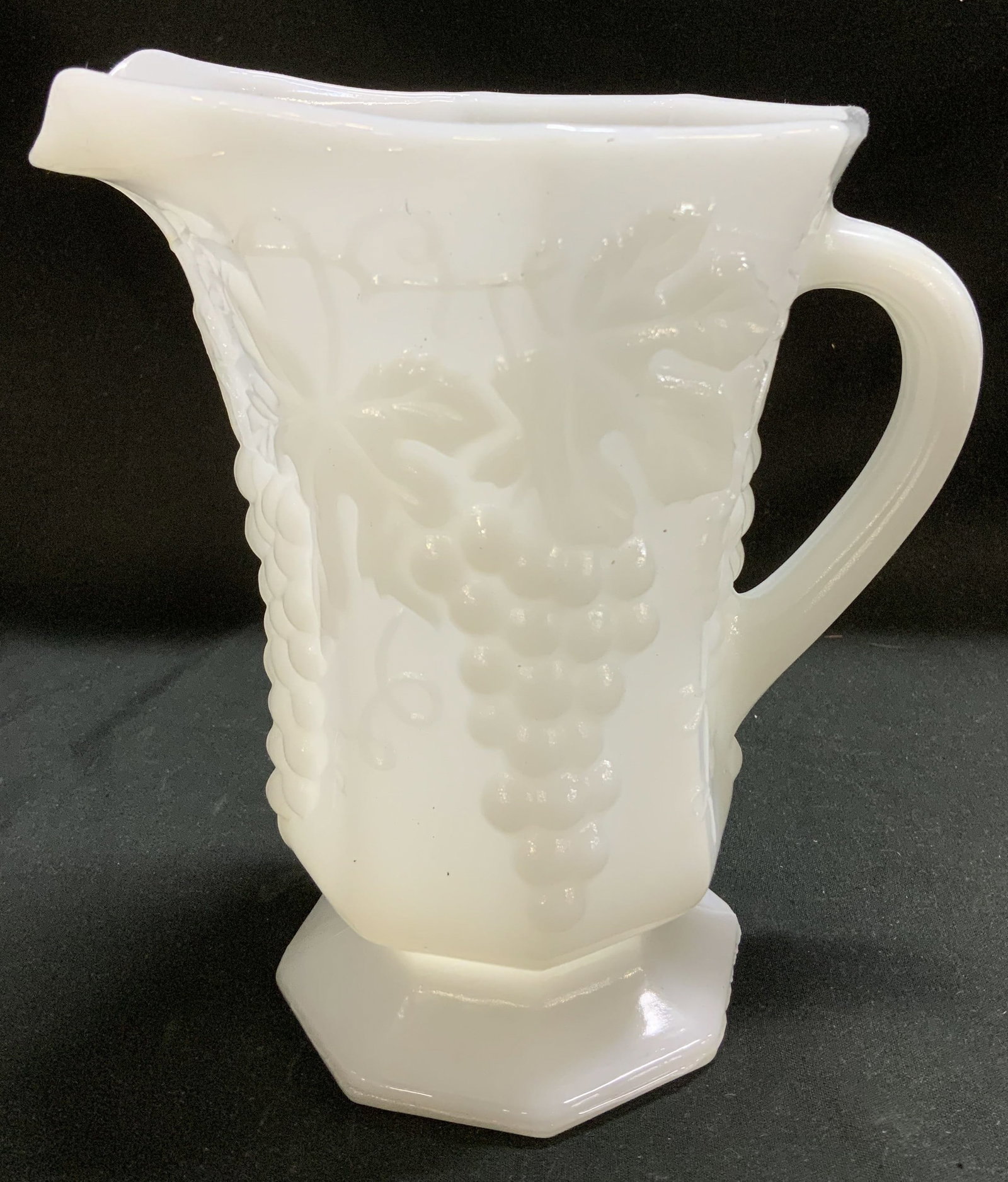 Anchor Hocking White Milk Glass Pitcher: Vintage Anchor Hocking white milk glass pitcher featuring a raised grapevine pattern. Footed base. Signed & marked on underside. Surface wear consistent to age, use & exposure. Measurements: length 5.