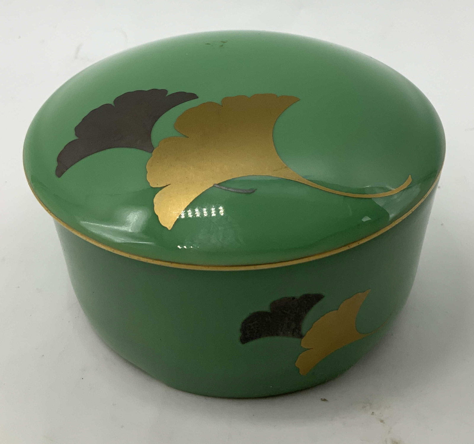 Sgnd Leaf Motif Porcelain Round Box: Porcelain round box is green toned & features gilt/dark leaf motifs on lid. Signed & marked on underside. Visible Asian characters. Surface wear consistent to age, use & exposure. Measurements: length