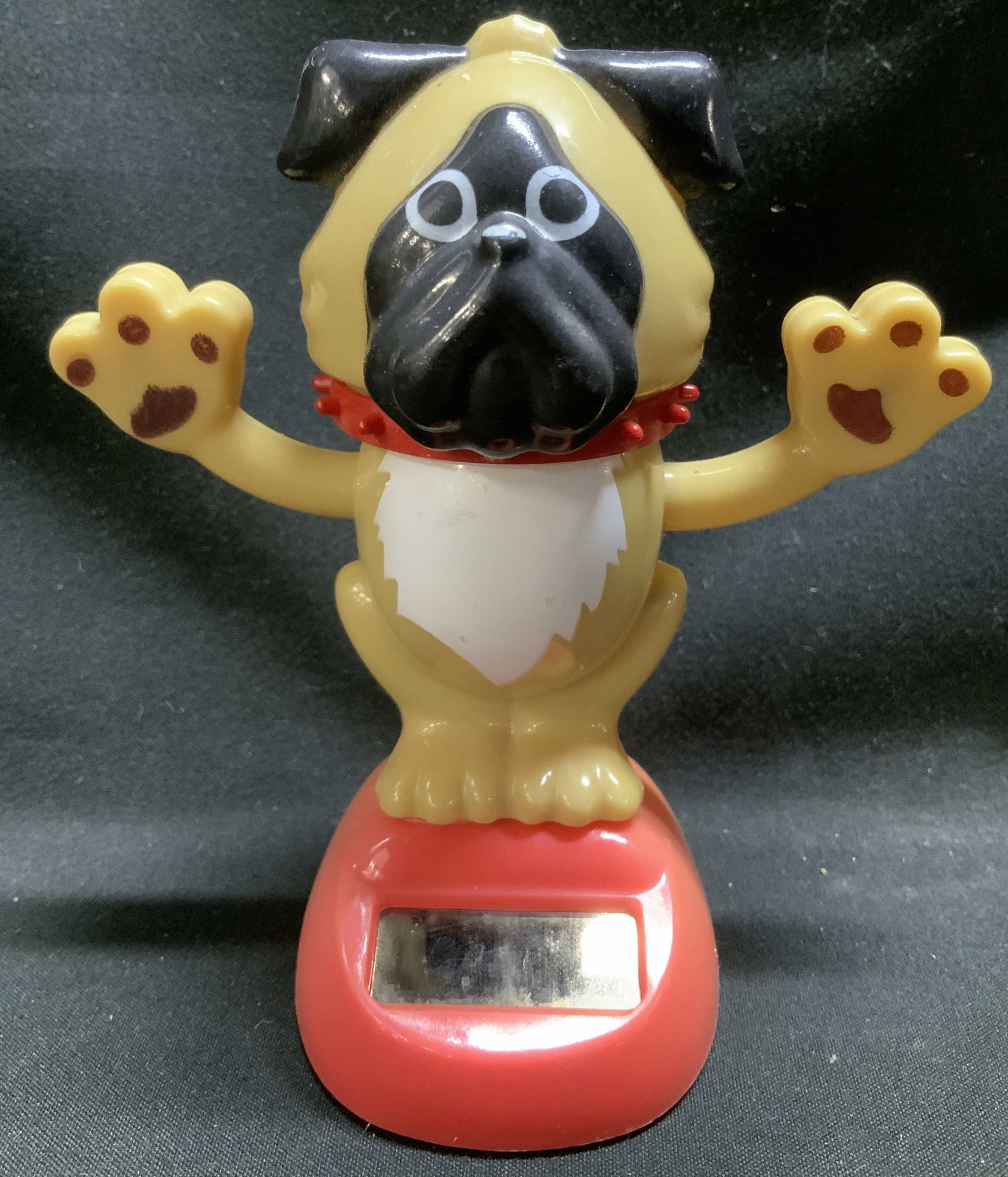 Solar Powered Pug Figurine 4.2in: Solar Powers figurine depicting a pug with its arm raised. Stands on base. Plastic material. No visible signature or markings. Surface wear consistent to age, use & exposure. Measurements: length of b