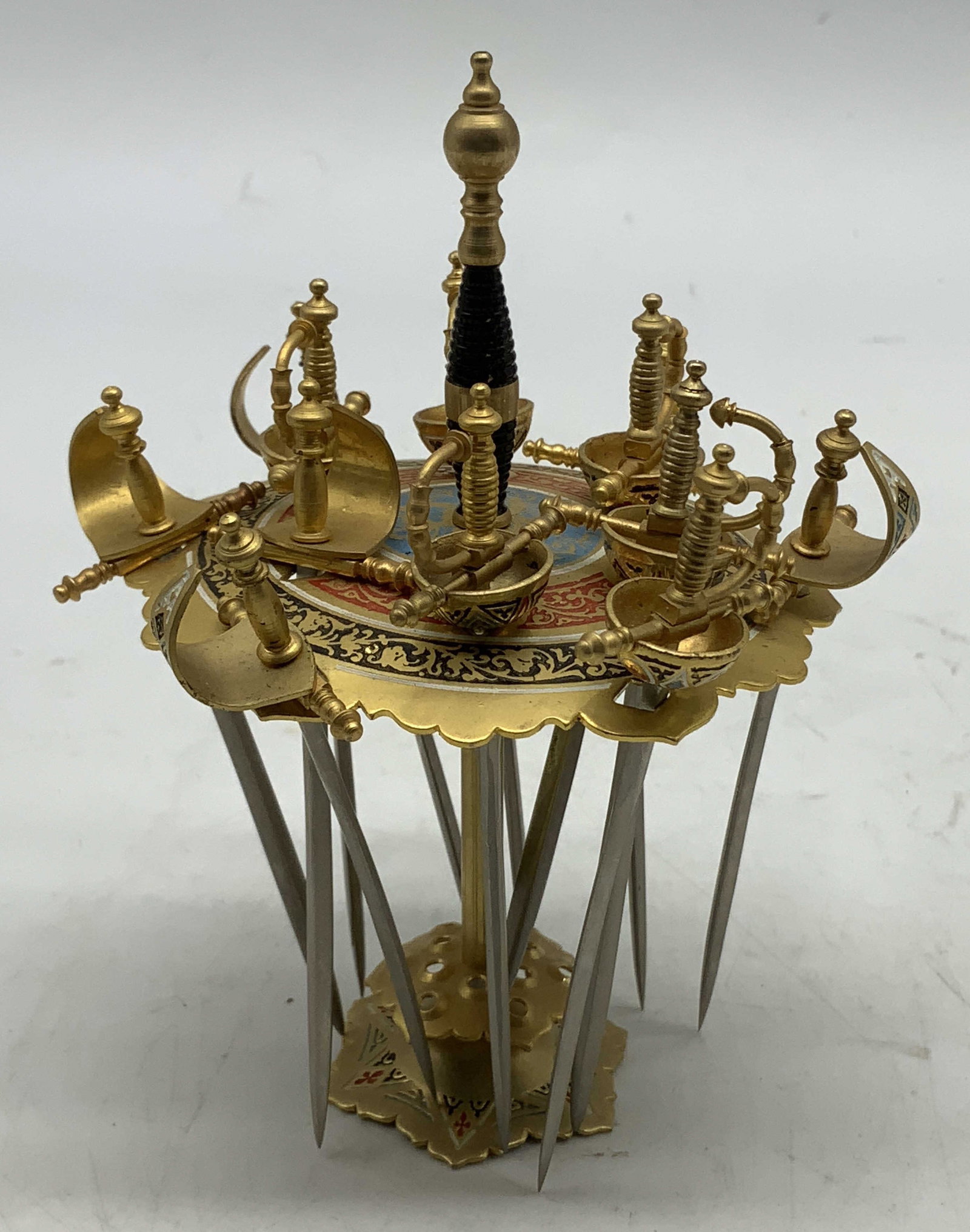 MCM Toledo Cocktail Sword Picks & Stand: Mid Century Modern cocktail sword picks with stand. 12 sword picks with intricate detailing on handles. Gold toned. Stand has blue, red & black scroll motifs. Surface wear consistent to age, use & exp