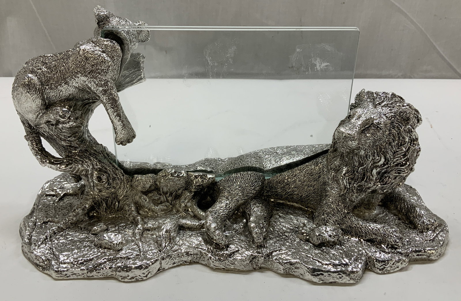 Silver Toned Lion & Cub Deco Photo Frame: Silver toned tabletop photo frame featuring a laying lion & perched cub. Rectangular glass panel. Textured finish. Silver toned metal. No visible signature or markings. Surface wear consistent to age,
