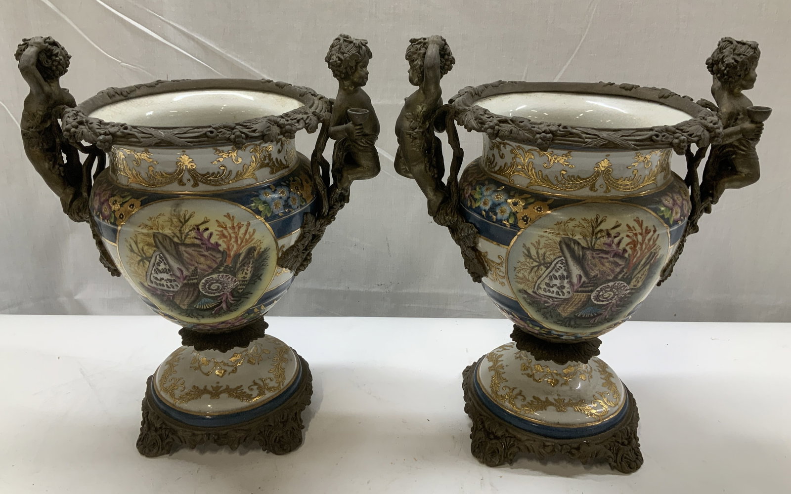 Pair Vtg Wong Lee 1895 Porc & Bronze Vase: Lot includes, a pair of vintage Wong Lee 1895 porcelain vases. Hand painted accents of marine shell & floral motifs. Gilt accents. Ornate bronze patina cherub & grapevine patterns. Pieces have weight.