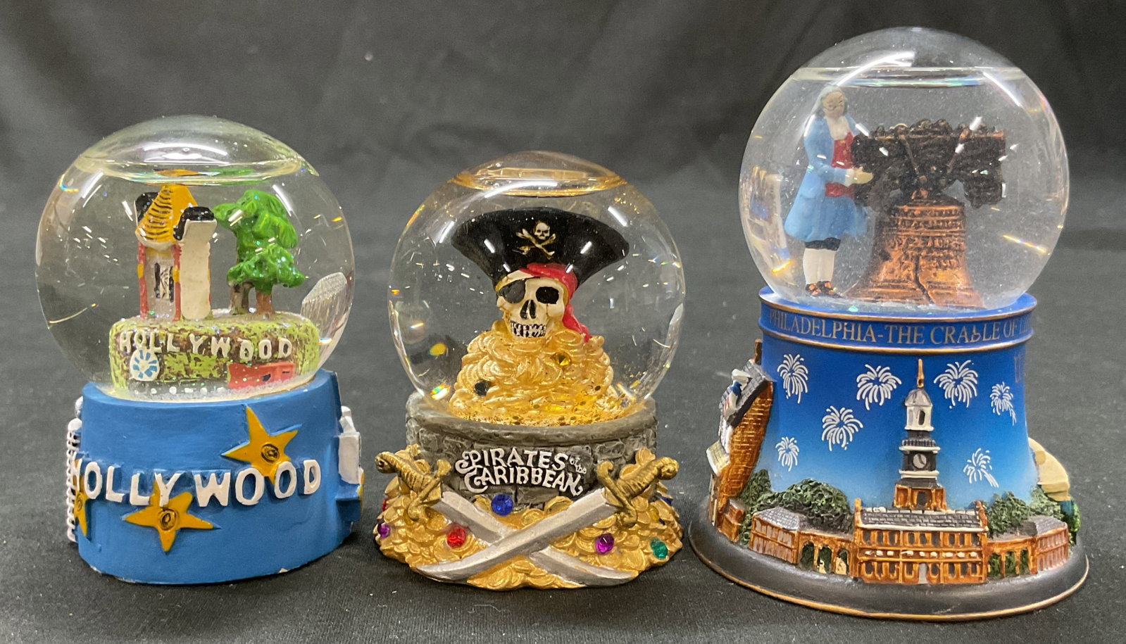Lot 3 Disney+ Souvenir Snow Globes: Lot of 3 souvenir snow globes. Includes Disney Pirates Of The Caribbean, Hollywood, and Philadelphia. Glass globes and resin bases. Hand painted details and features. Varying colors. Largest measures 