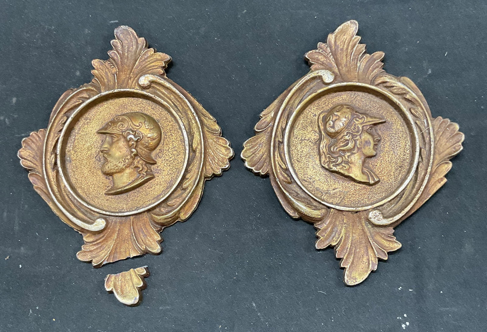 Pair Bronze Patina Roman Style Wall Plaques: Pair of vintage bronze patina metal wall plaques. Ornate scroll frames with Roman style cameos. Measures approx. 6.5 x 5 inches. Wear including patina and scratching. One piece needs repair. Bronze