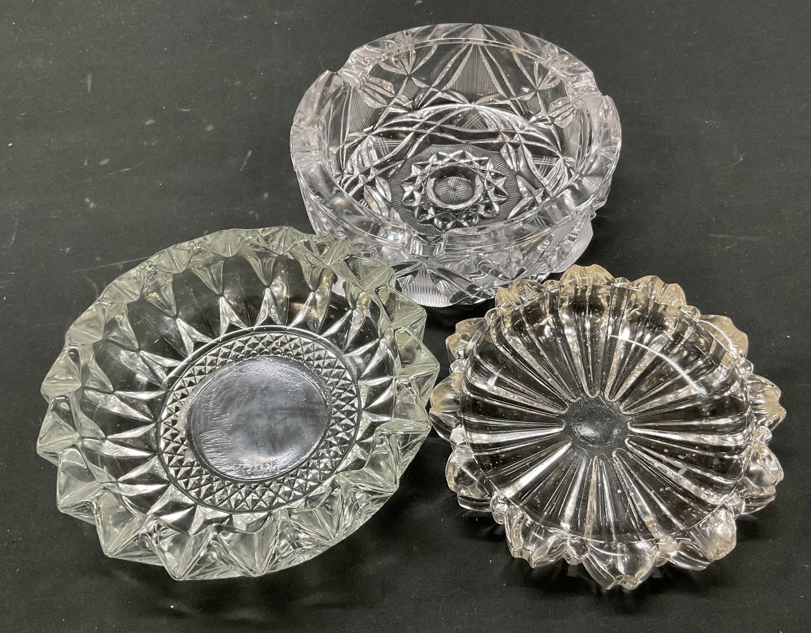 Lot 3 Vintage KIG+ Glass Ashtrays: Lot of 3 vintage glass ashtrays. One is marked KIG. Round ashtrays. Scalloped and sawtooth edges. Cut geometric patterns. Textures surface. Indentations on rim. Largest measures ht approx 2 inches dia