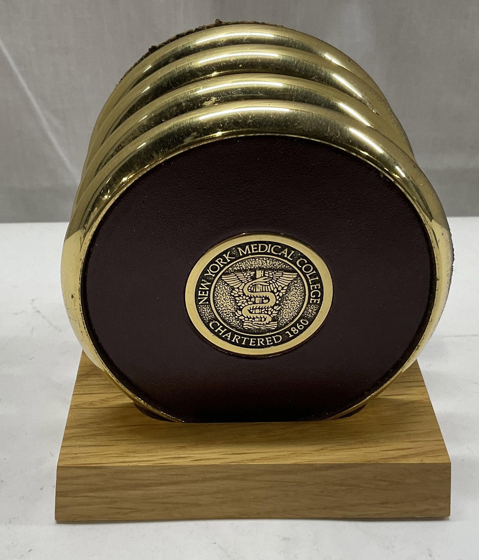 St 4 New York Medical College Coasters: Set of 4 New York Medical College Coasters. Gold-Toned Metal Rims and College Seal in the Middle. Reddish-Brown Leather-Style Center. Comes in Wooden Stand. Condition Consistent with Exposure. Measure