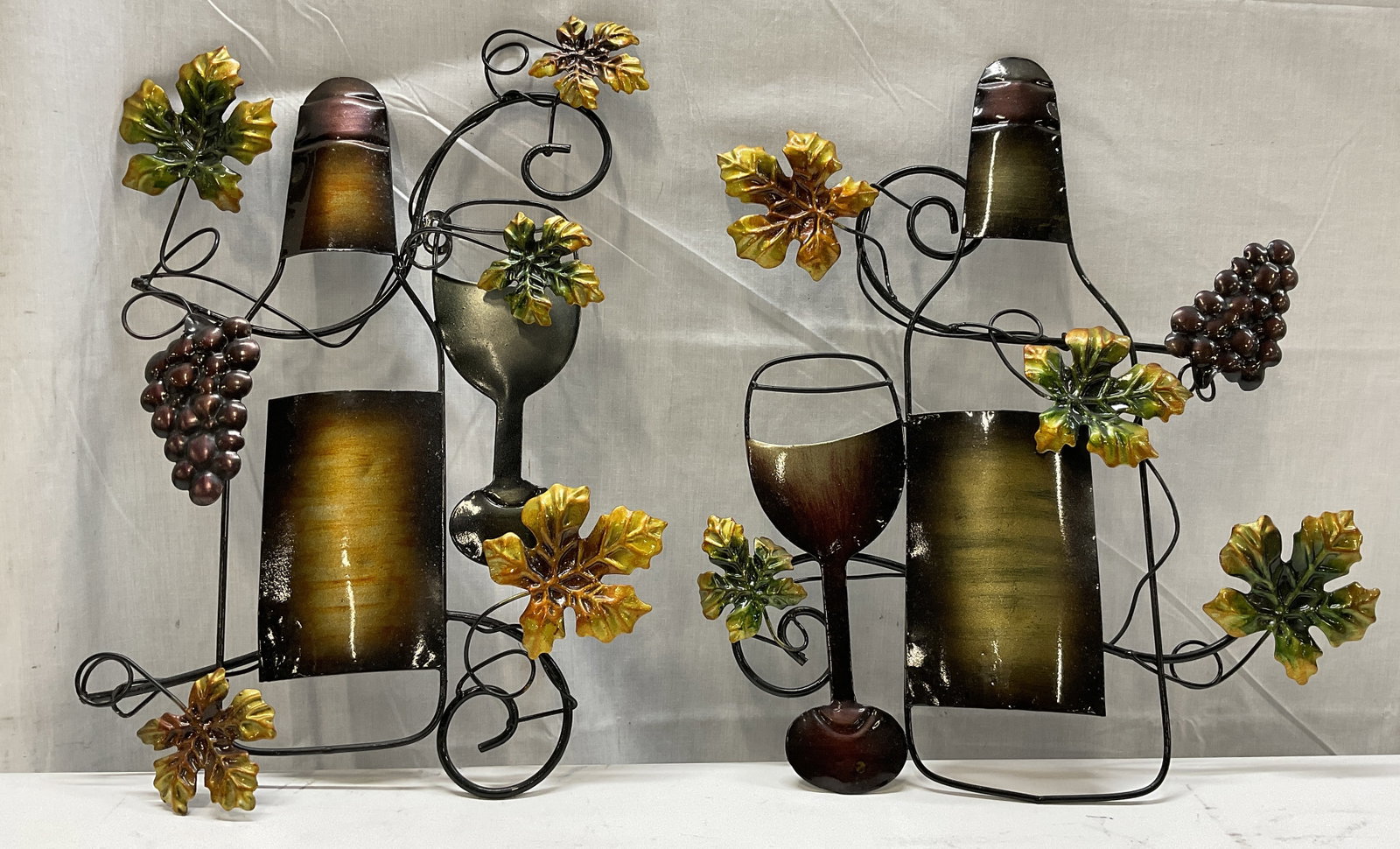 Set 2 Metal Wine Bottle 17.5in Wall Art: Set of 2 Metal Wine Bottle Wall Art. Made of Steel with Varying Painted Earth Tones on Each. Features 2 Wine Bottles with Glasses Wrapped in Scrollwork and a Grapevine Design. Condition consistent wit