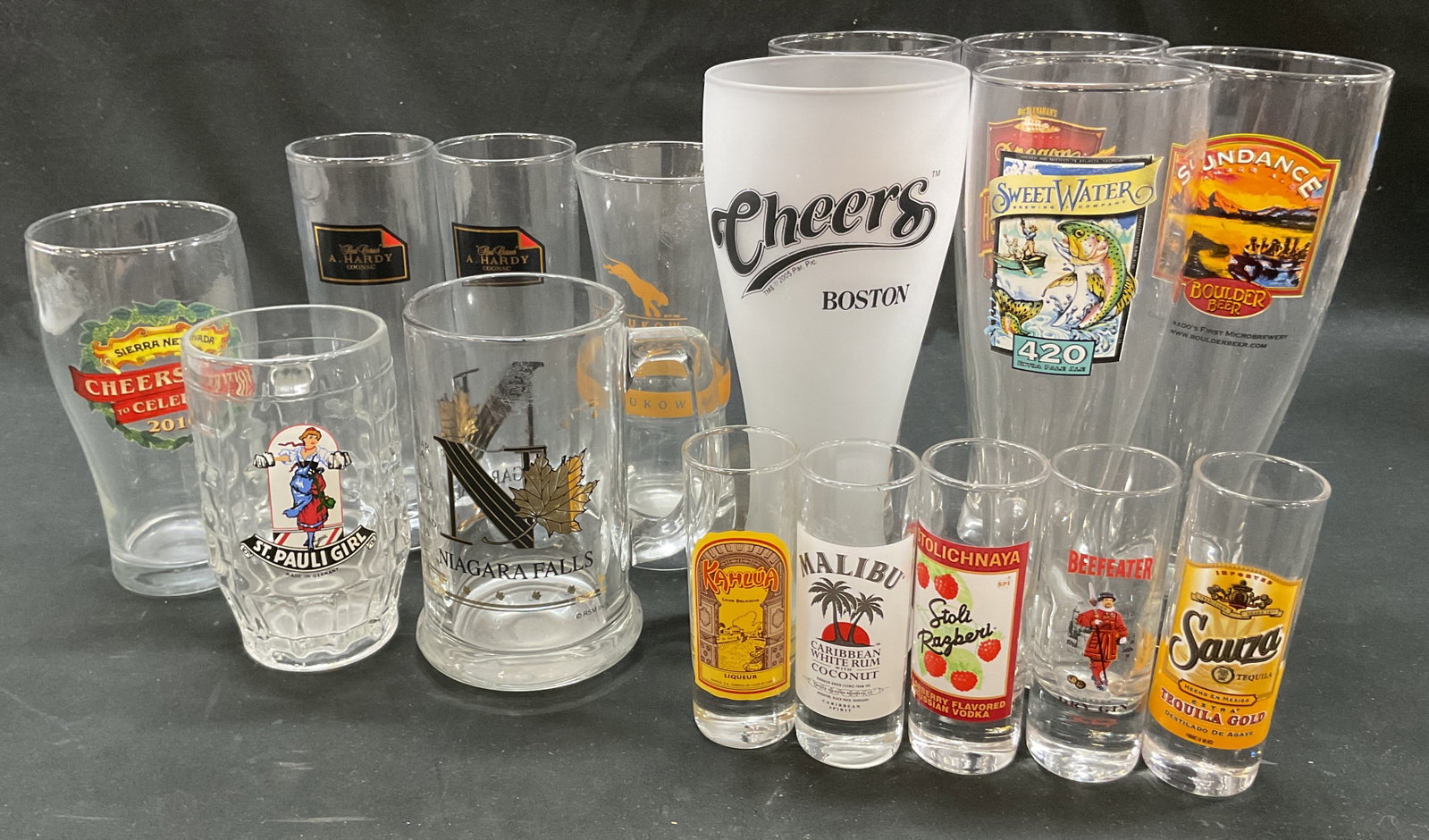 Grp Lot 16 Assorted Glass Barware: Group lot of 16 assorted glass barware. Includes beer glasses, shot glasses, beer mugs, and more. Varying designs and colors. Some include Oregon Honeybeer, Beefeater, Malibu, Niagara Falls, and more.