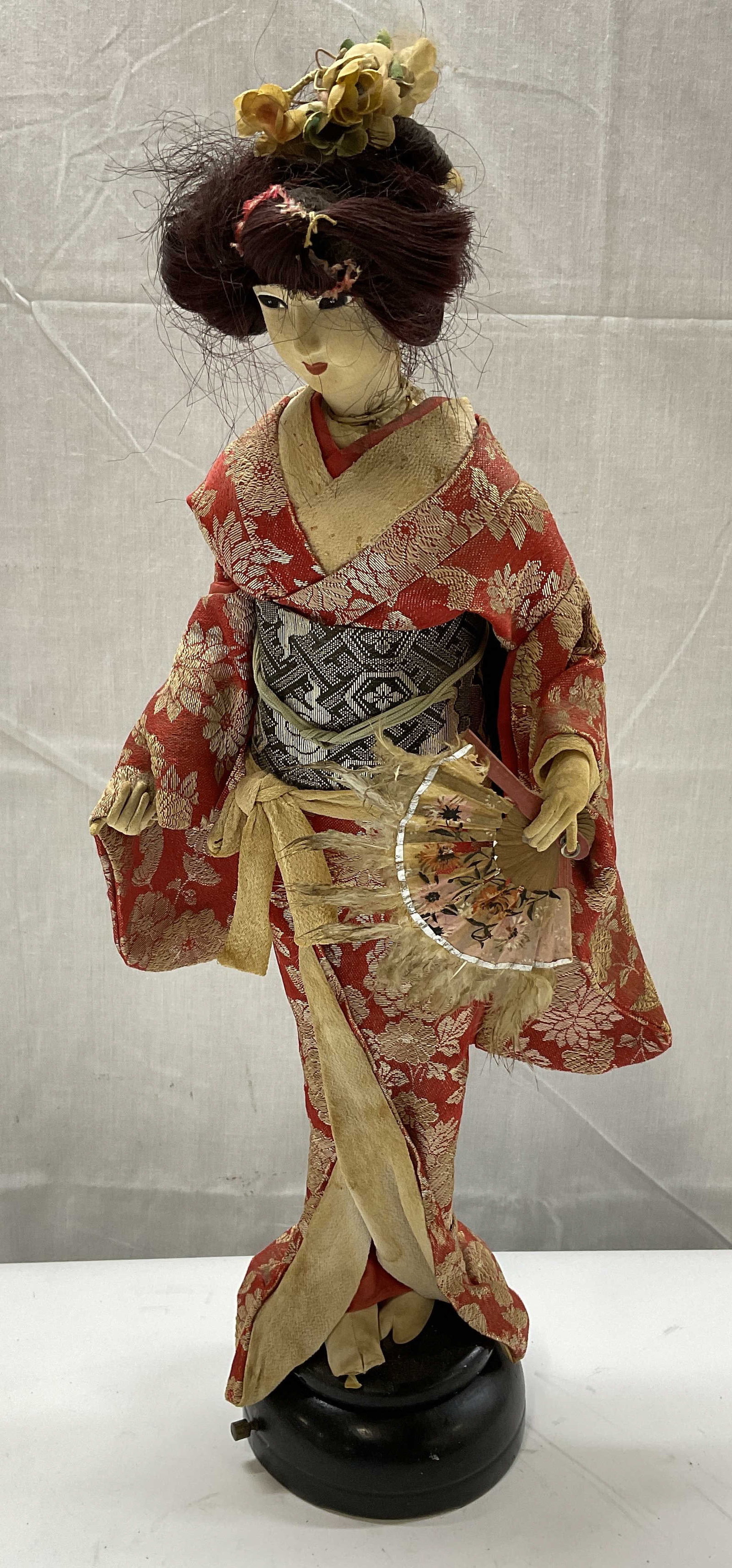 VTG Japanese Geisha Doll: Vintage Japanese Geisha Doll in Red Floral Kimono on Musical Stand. Body Made of Fabric. Holds Paper Fan with Feather Accents. Not Tested for Functionality. Measures app 19.5in H x 4.5in Base. Surface