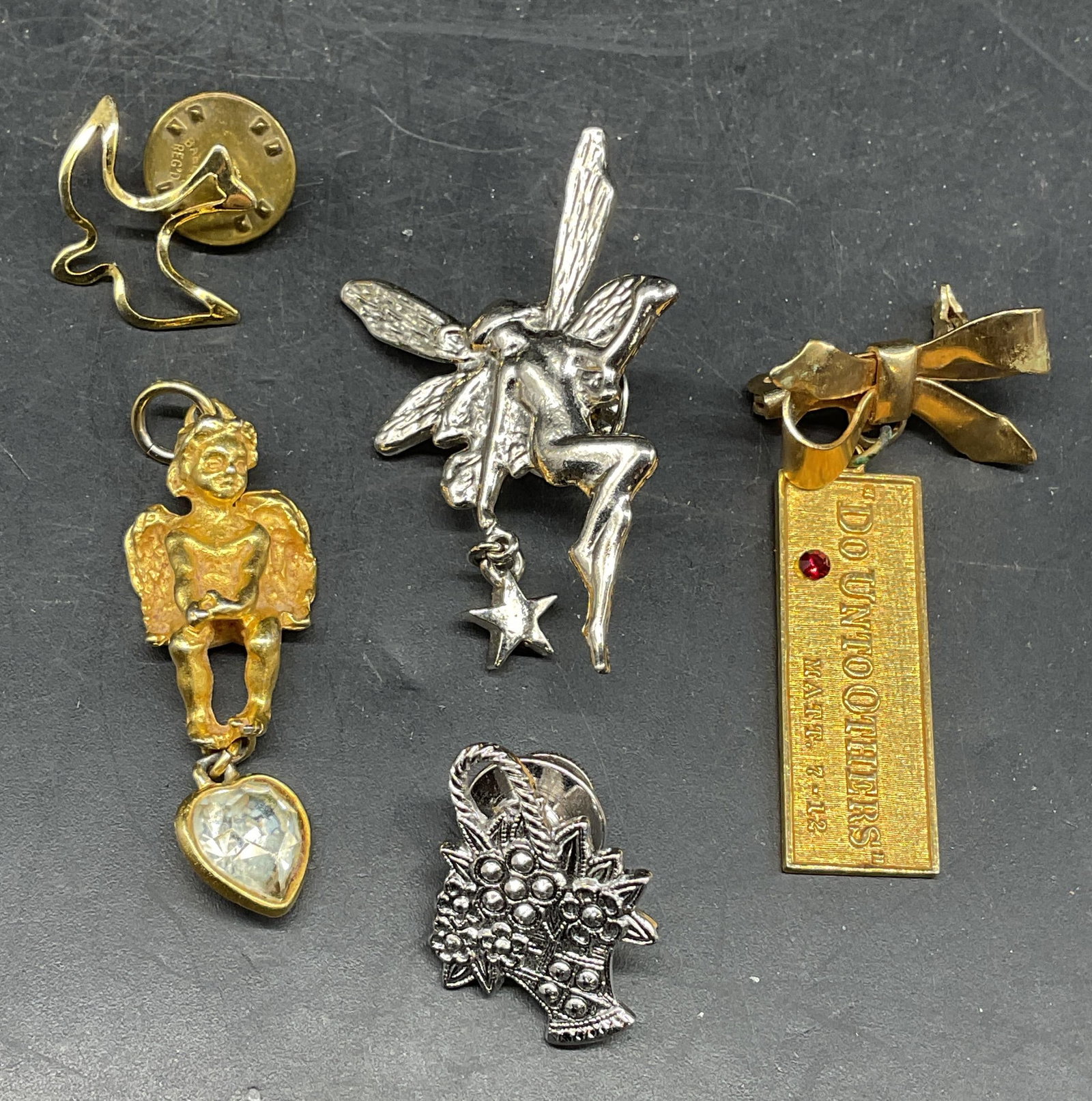Lot 5 Avon Flower Basket Pin, Cherub + More: Lot of 5 gold and silver tone pins, brooch, and pendant. Signed Avon silver tone flower basket pin, silver tone fairy pin, gold tone dove pin, gold tone cherub pendant, and gold tone religious motif a