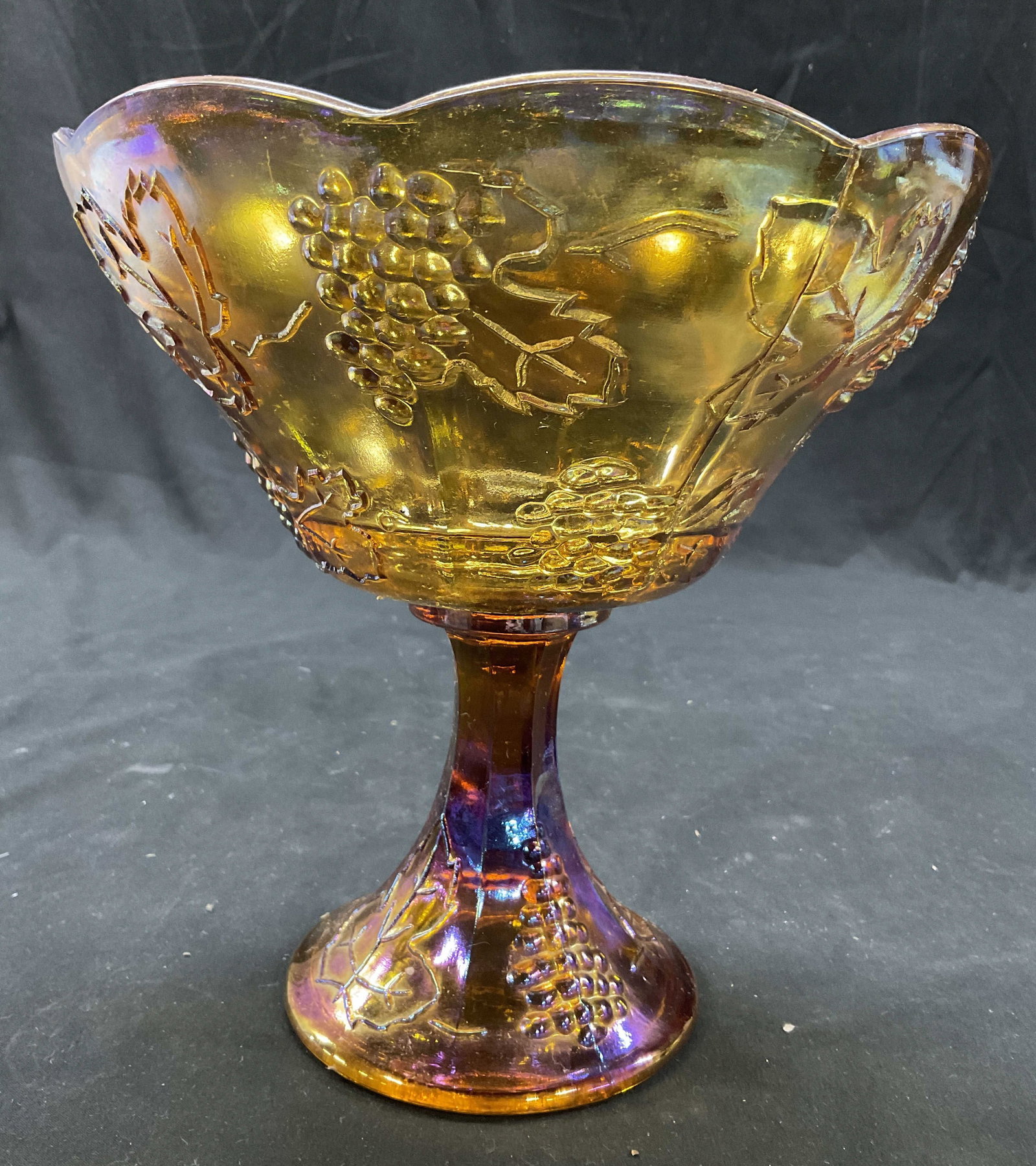 Carnival Glass Indiana Glass footed Compote Bowl: Vintage collectible Harvest Grape Pattern Amber Carnival Glass, molded design of grapes and leaves on bowl and base, iridescent finish creates multi colored reflections, fluted scalloped top rim,