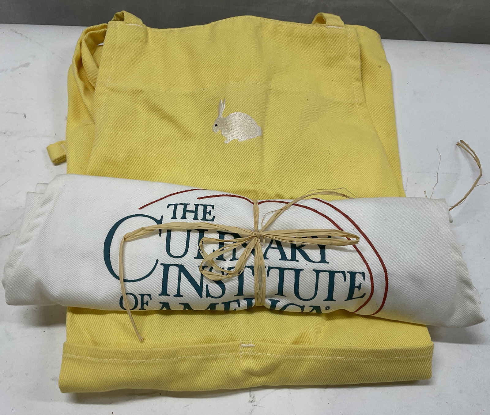 Lt 2 Cotton Aprons CIA+: 2 Cotton Aprons. White Culinary Institute of America Apron Tied with Twine and a Yellow Apron with an Embroidered Rabbit Made in Vietnam. Condition Consistent with Exposure. Measures app 24.5in L x