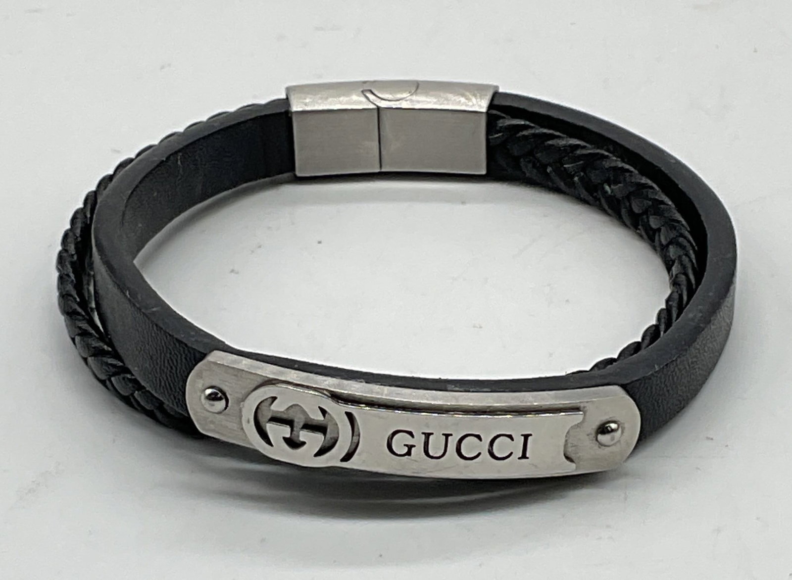 Gucci Black Leather Silver Tone Monogram Bracelet: Gucci black leather solid and braided strap bracelet with silver tone metal monogram plaque. Measures approx. 7.5 inches long. Some scratching. Jewelry, estate jewelry, costume jewelry, cocktail