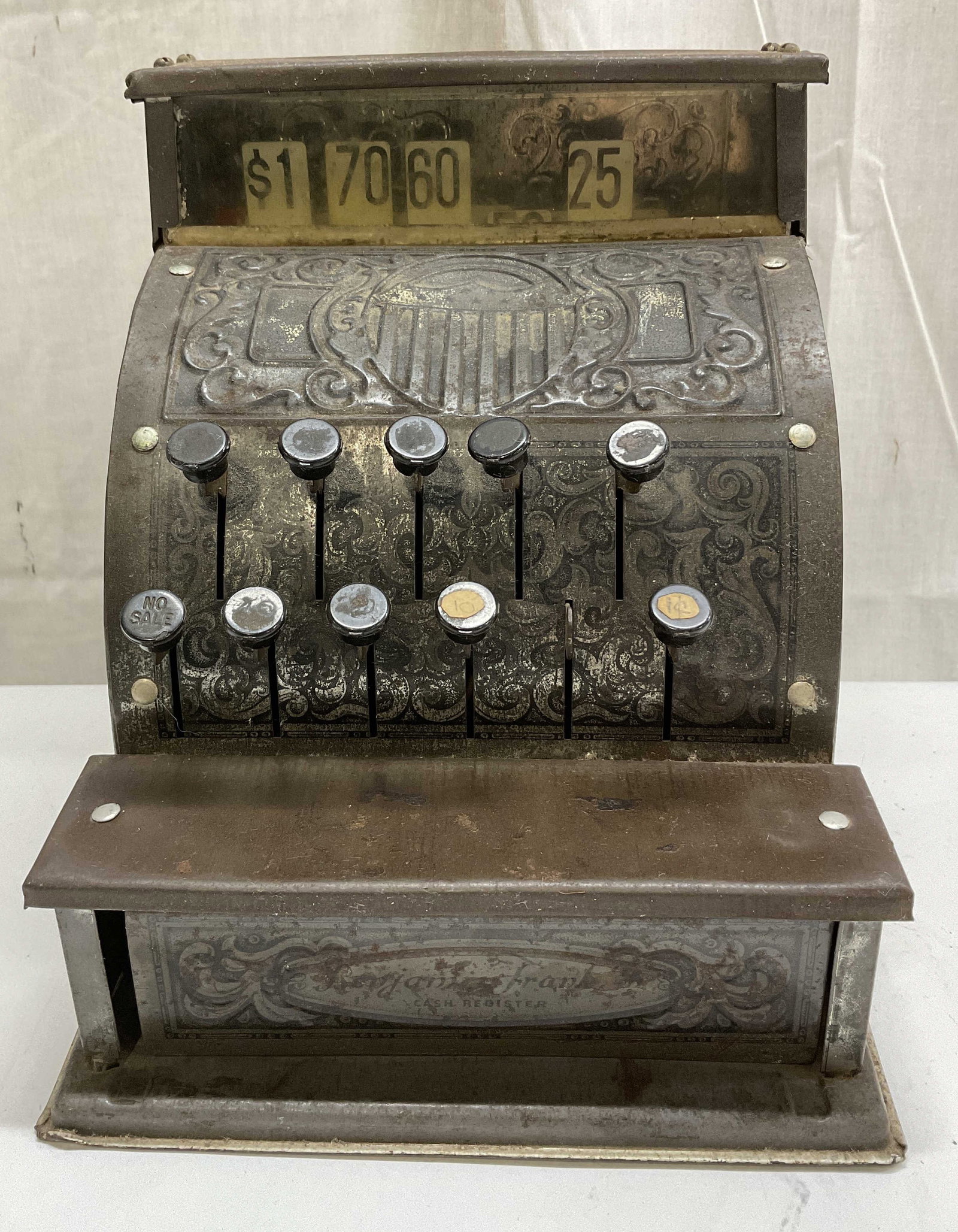 VTG Benjamin Franklin Tin Litho Cash Register: Vintage Benjamin Franklin Tin Litho Cash Register. Floral and Shield Designs. Aging to Metal, Scratching, and Bending Present. Button Missing. Not Tested For Functionality. Measures app 7.5in H x 6.75