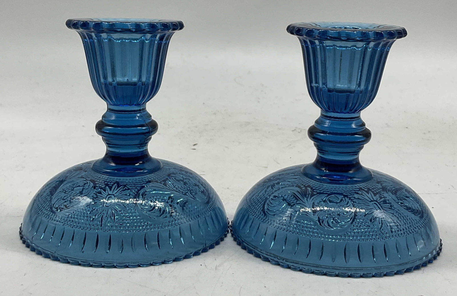 Pair Vintage Blue Glass Candlestick Holders: Pair of vintage blue glass candlestick holders. Round scalloped base. Ornate floral and scroll motifs. Ribbed surface. Bulb like stem. Scalloped edge top. Measures ht approx 4 inches diam approx 3.5 i