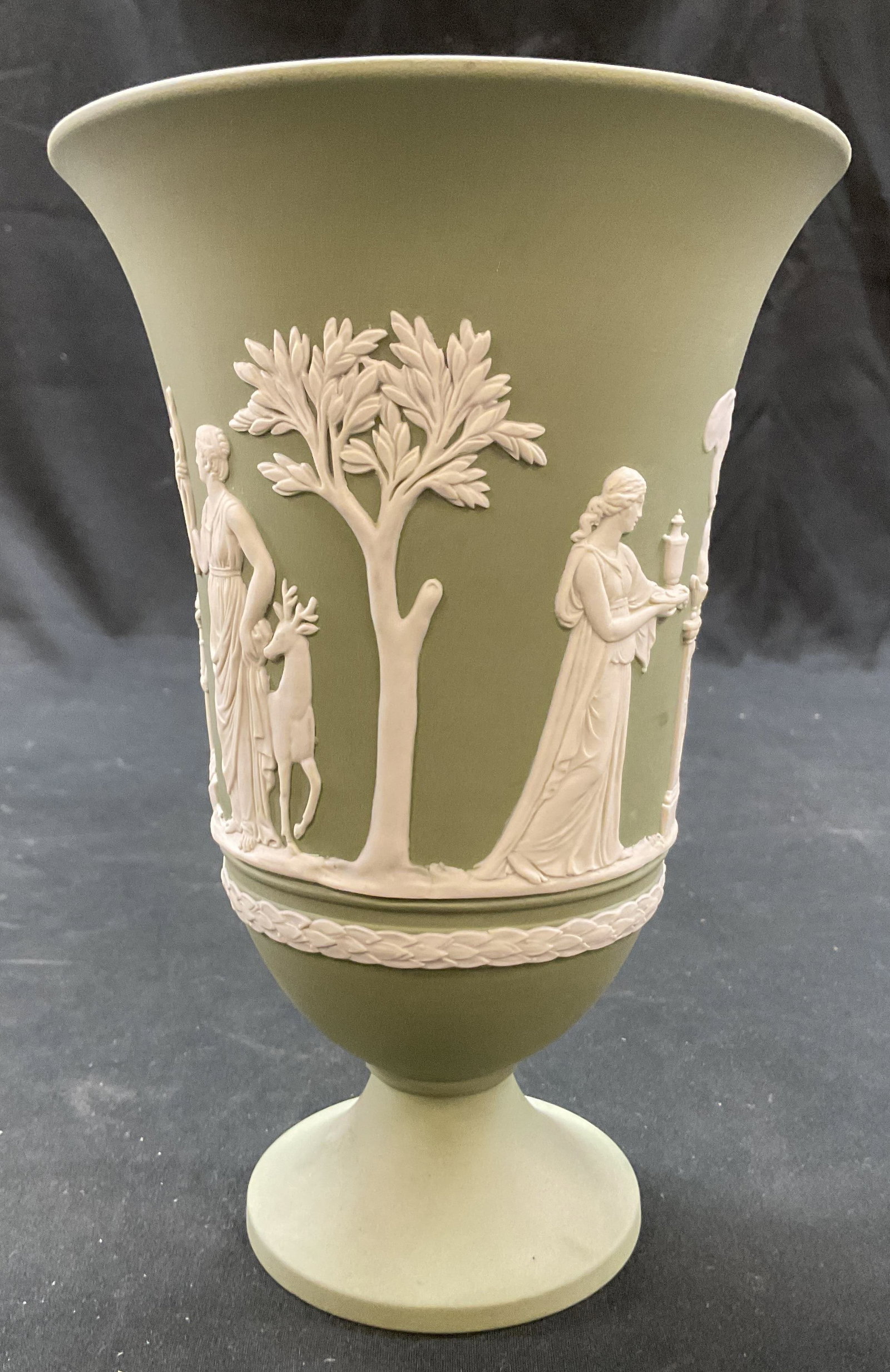 Wedgwood Green Jasperware Vase England: Wedgwood green Jasperware vase. Marked on underside, reads Wedgwood Made In England. Round pedestal base. Wide rim. White bas relief neoclassical decorations. Figural and leaf motifs. Measures ht appr