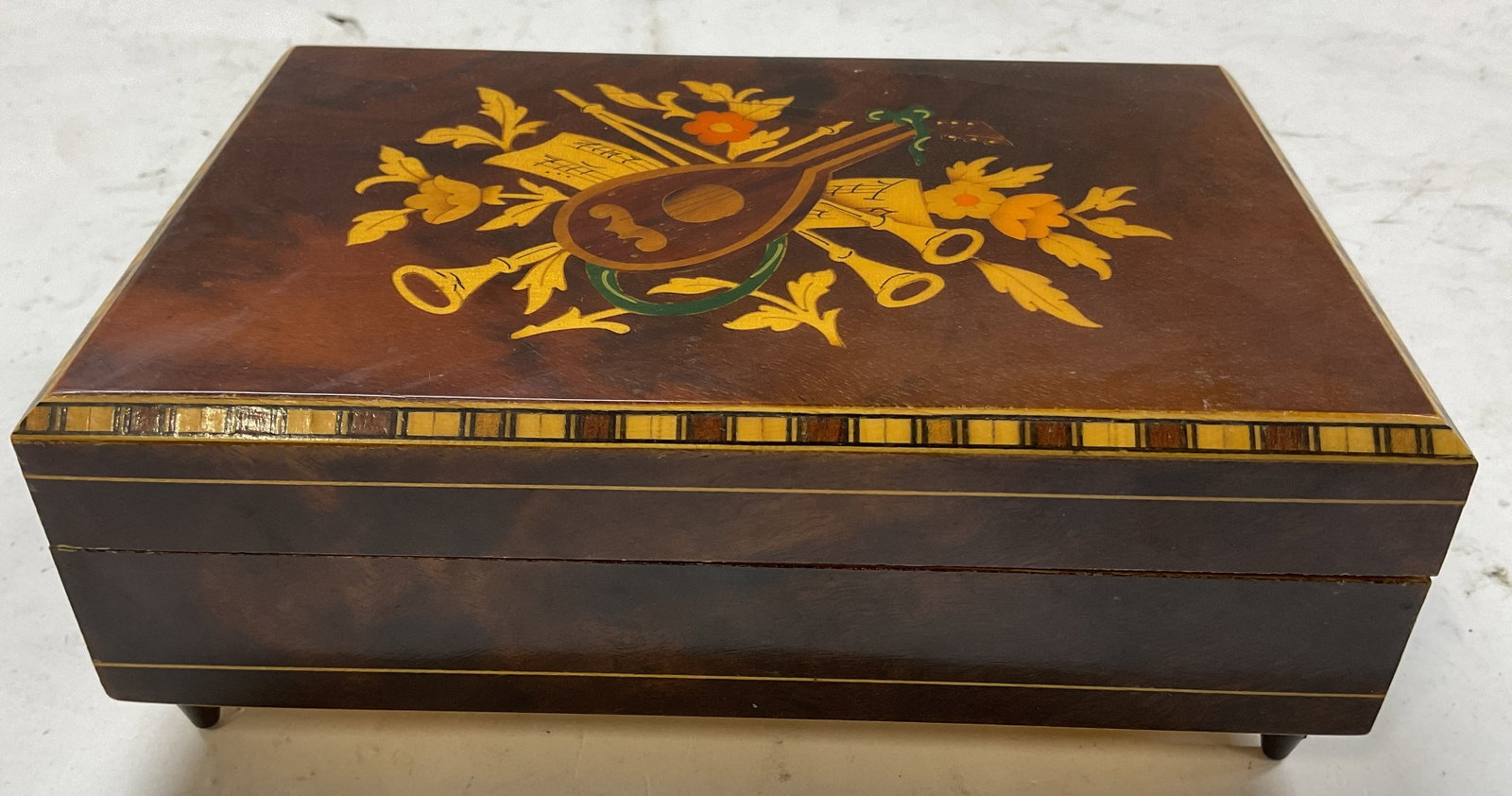 Vintage Wood Inlay Footed Music Jewelry Box Italy: Vintage wood inlay footed music jewelry box. Made in Italy. Four feet on underside. Rectangular shape. Inlaid floral, scroll, and musical instruments motifs in varying colors. Decorative border.