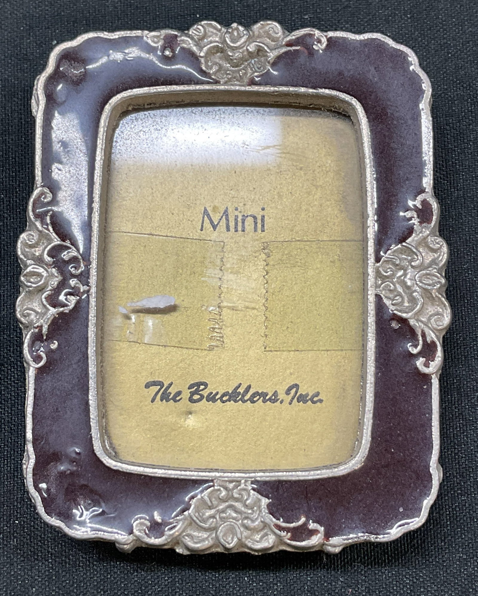 The Bucklers Inc Silver Tned Enamel Picture Frame: The Bucklers Inc silver toned enameled picture frame. Marked The Bucklers Inc. Silver toned floral motifs with red enameled inlay. Scalloped edge. Measures approx 2 x 2.5 inches. Surface wear
