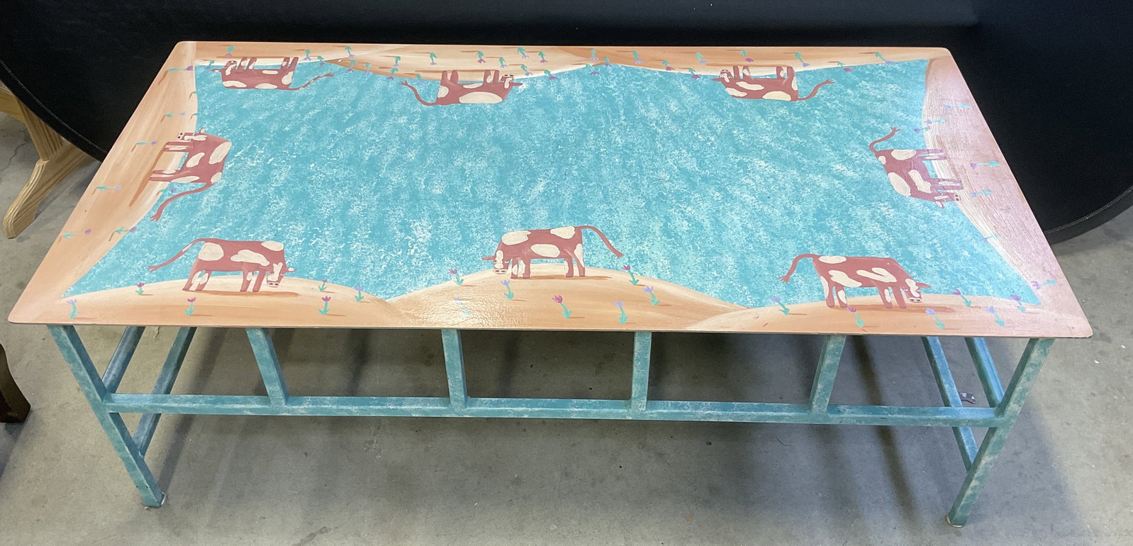 Southwestern-Style Painted Cow Motif Iron Table: Southwestern-Style Iron Table. Folk Art Style. Painted Cow Motifs on Hills with Florals and a Speckled Turquoise Pond Center. Condition Consistent With Exposure. Measures app 16.5in H x 48in L x 24in 