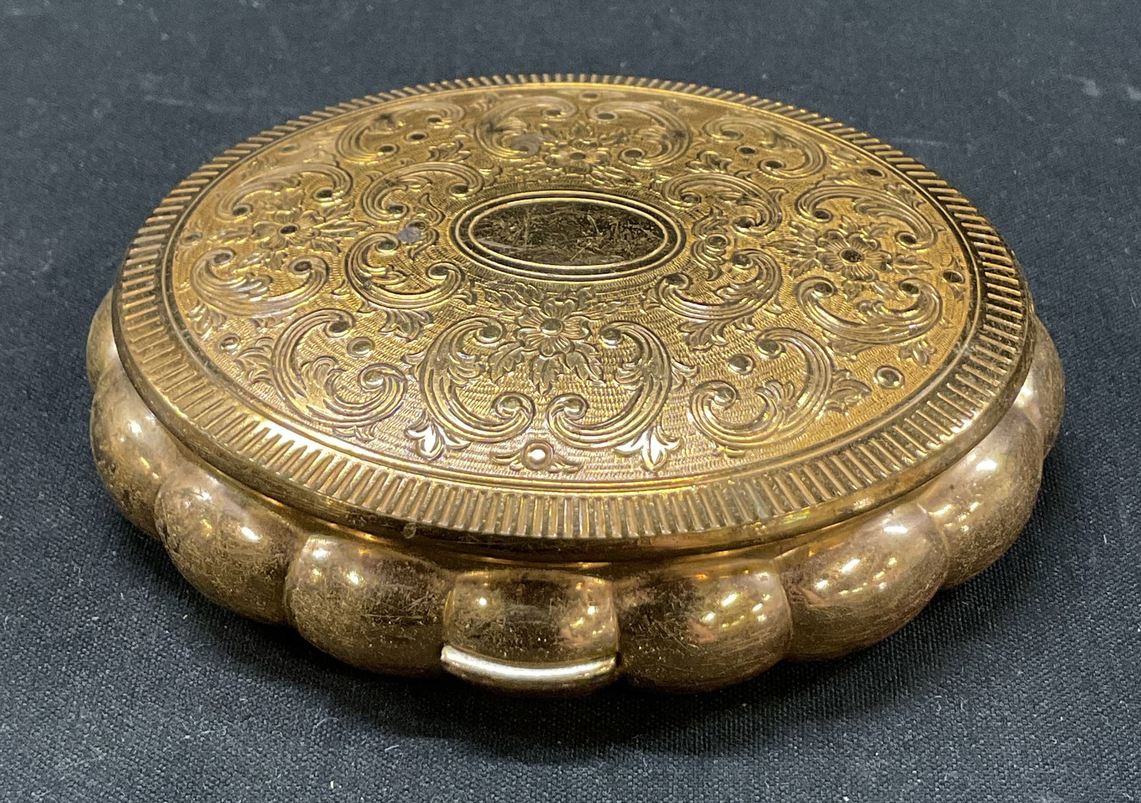 Kigu Minuette Musical Powder Compact England: Kigu Minuette gold toned metal hinged musical powder compact. Marked on surface. Made in England. Oval shaped. Scalloped edge. Hinged lid. Embossed scroll and floral motifs. Fitted compartments and mi