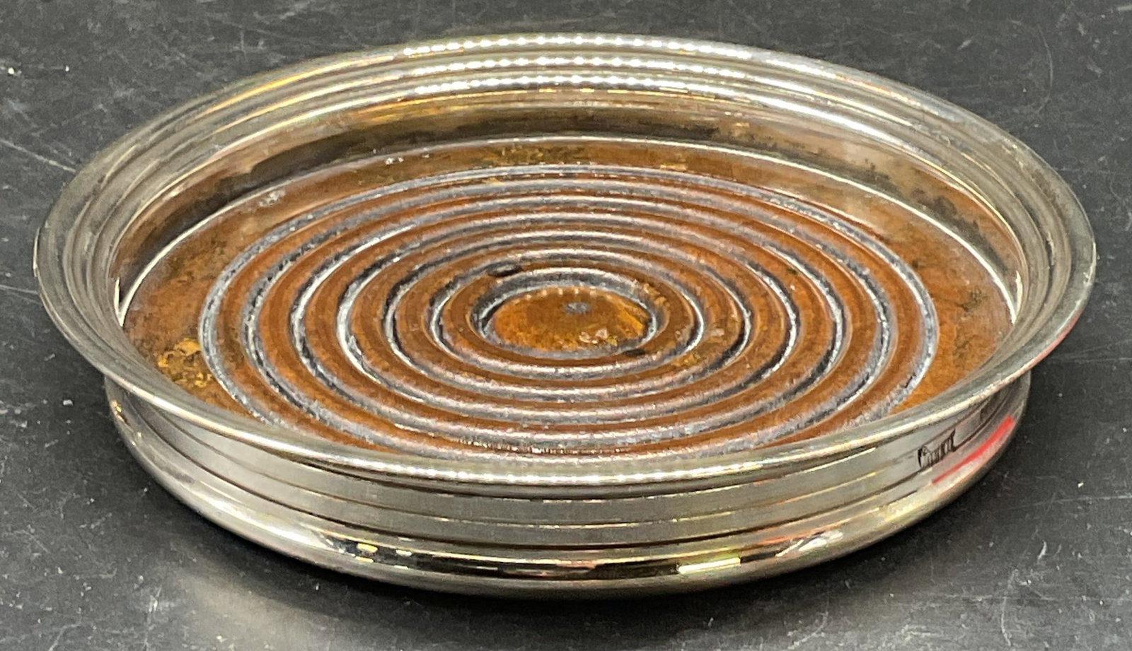 Signed Silver Plated Bottle Coaster, Spain: Signed silver plated bottle coaster. Made in Spain, signed illegibly. Wood base. Measures approx. 5 x .75 inches. Wear including scratching, loss to plating. Silver plated bottle coaster, bottle coast