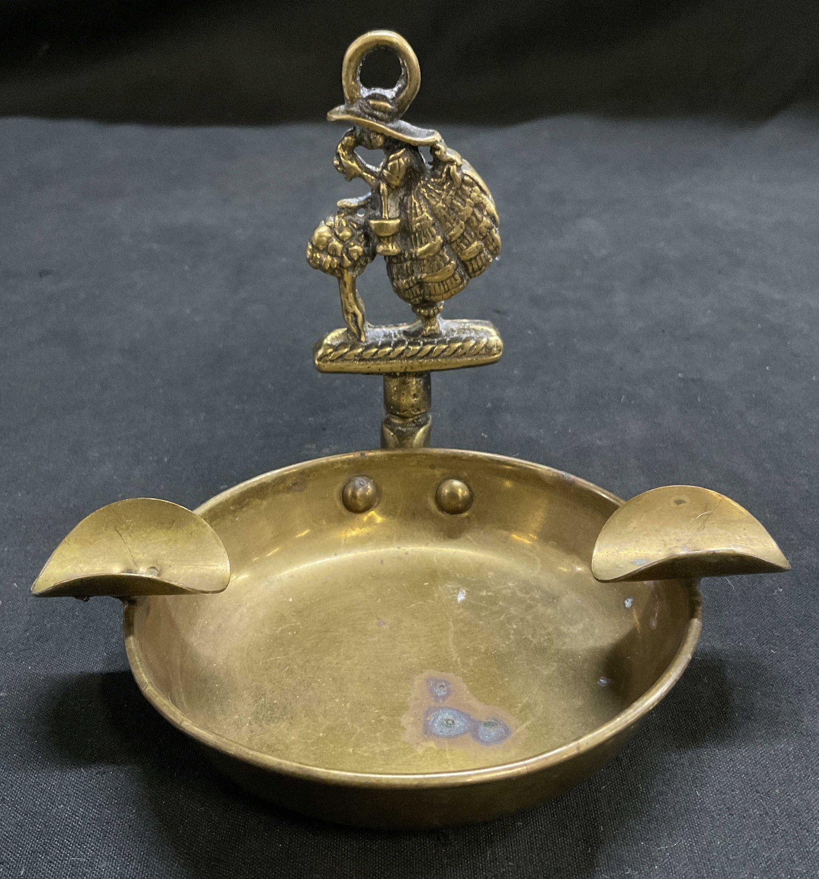 VINTAGE Colonial Woman Figural Brass Ashtray: Vintage Brass Ashtray. Colonial Woman in Large Dress Smelling Flowers Figure at Top. Two Cigarette/Cigar Rests. Scratching and Oxidization Present. Measures app 4in H x 4.75in Diam. Vintage Ashtray,