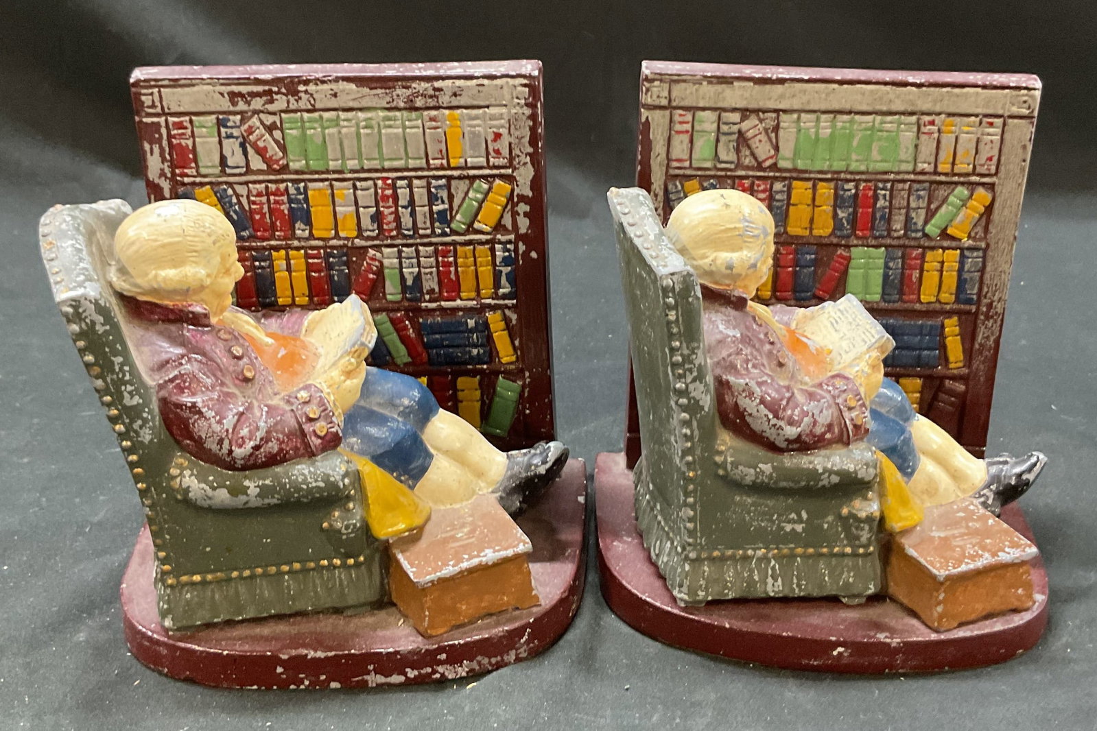 Pair Vtg Cast Metal Colonial Man Reading Bookends: Pair of vintage cast metal colonial man reading bookends. These bookends depict a colonial era man sitting in a wingback chair with his feet up reading a book. He sits next to a bookshelf filled with