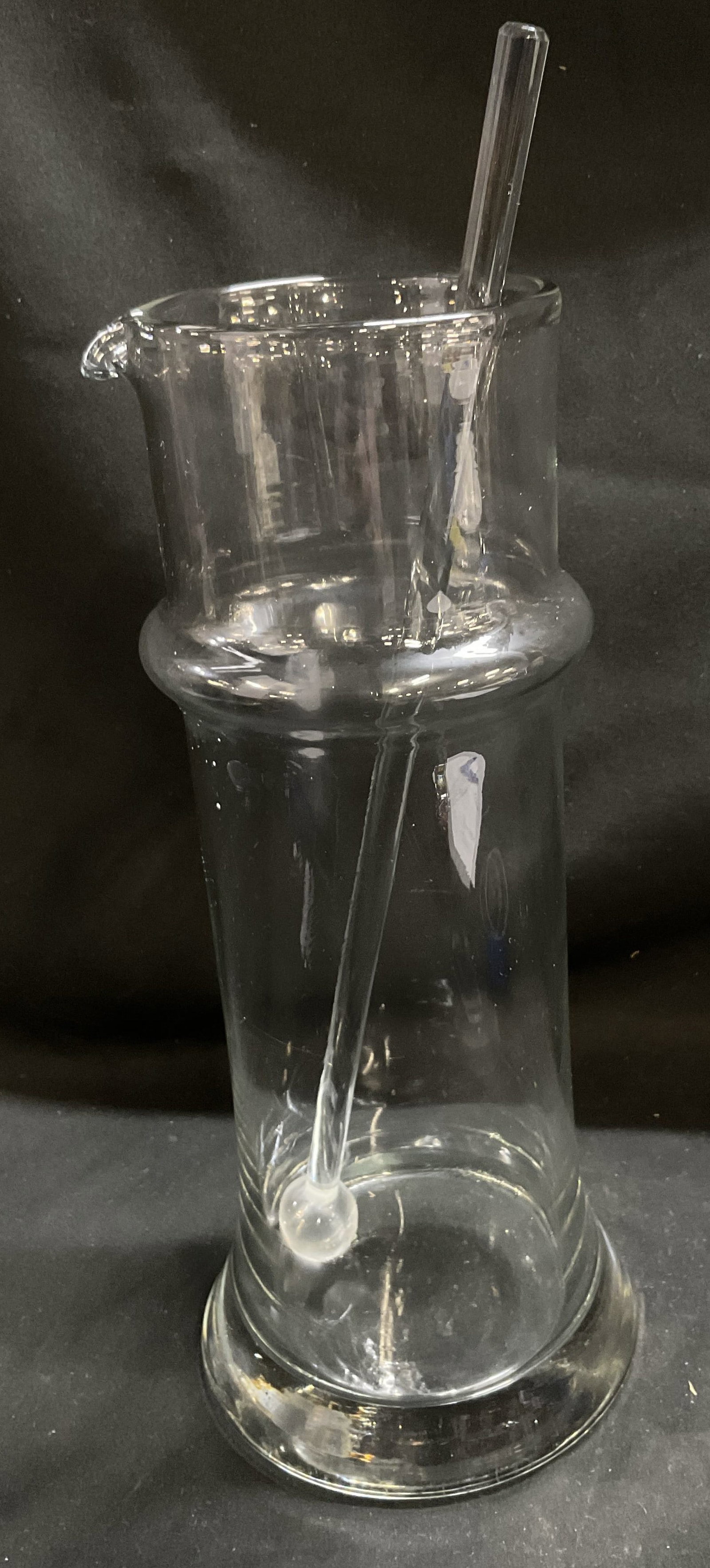VTG Clear Glass Martini Pitcher and Stirrer: Vintage Clear Glass Martini Pitcher and Stirrer. Minimalist Silhouette. Raised Ring on Upper Body. Slightly Flared Base. Frosted Ball Tip on Stirrer. Some Scratching. Measures app 12in H x 5in Diam. V