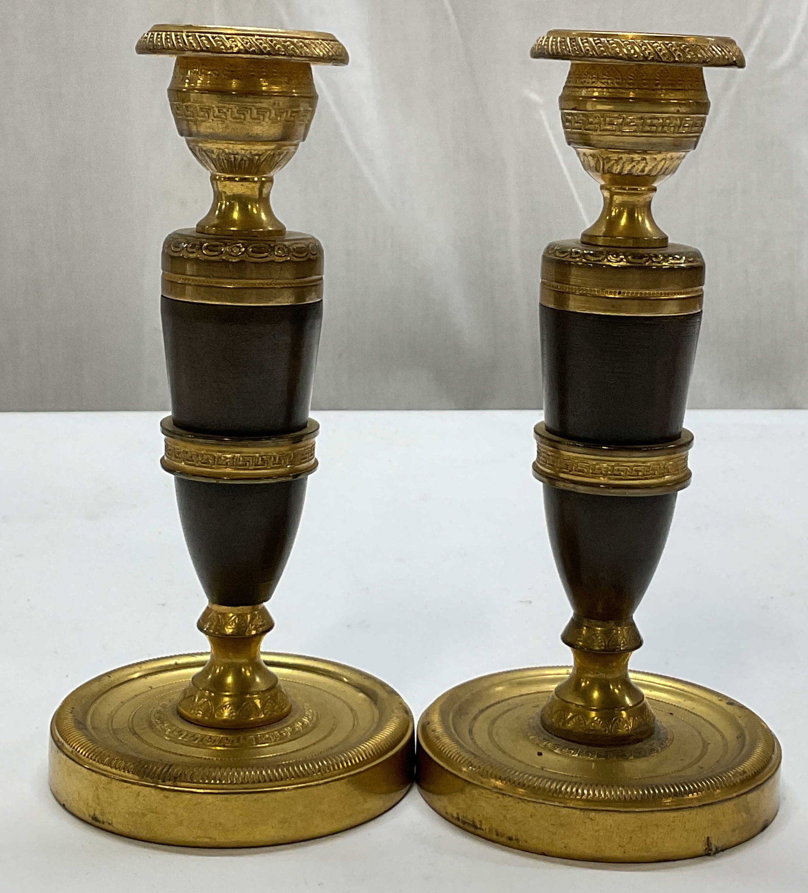 Pair Antq French Empire Style Brass Candlesticks: Pair of antique French Empire style brass candlesticks. Round base with ribbed trim. Urn shaped top. Multi tiered tapered main pillar. Embossed neoclassical motifs. Dark patina accent. Measures ht app