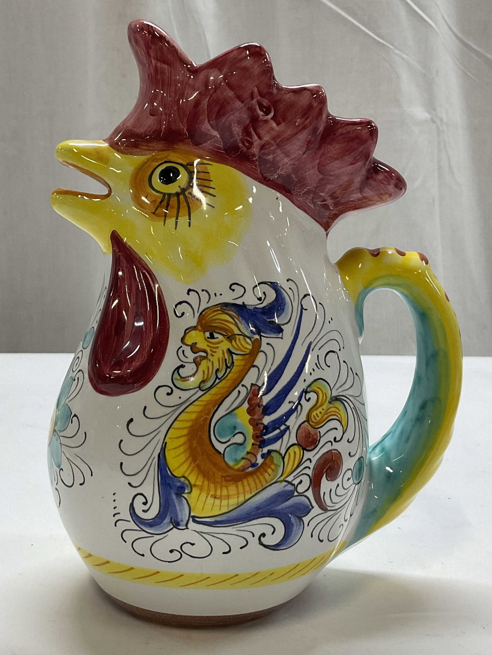 Williams Sonoma Ceramic Rooster Pitcher Italy: Williams Sonoma ceramic rooster pitcher. Marked on underside, made in Italy. Scalloped edge. Curved handle and spout is the mouth of the rooster. Hand painted features with floral and animal motifs. V
