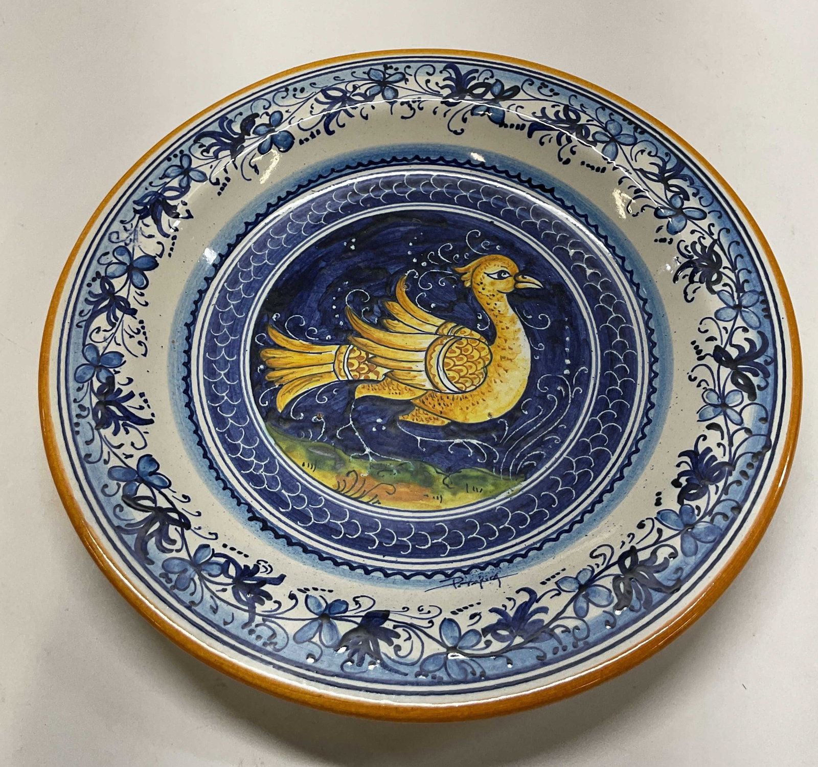Ceramiche Marino Hand Painted Wall Plate Italy: Ceramiche Marino hand painted ceramic wall plate. Marked on underside, made in Italy. Hook on underside for hanging. Orange trim. Central bird figure. Hand painted blue and white floral and scroll mot
