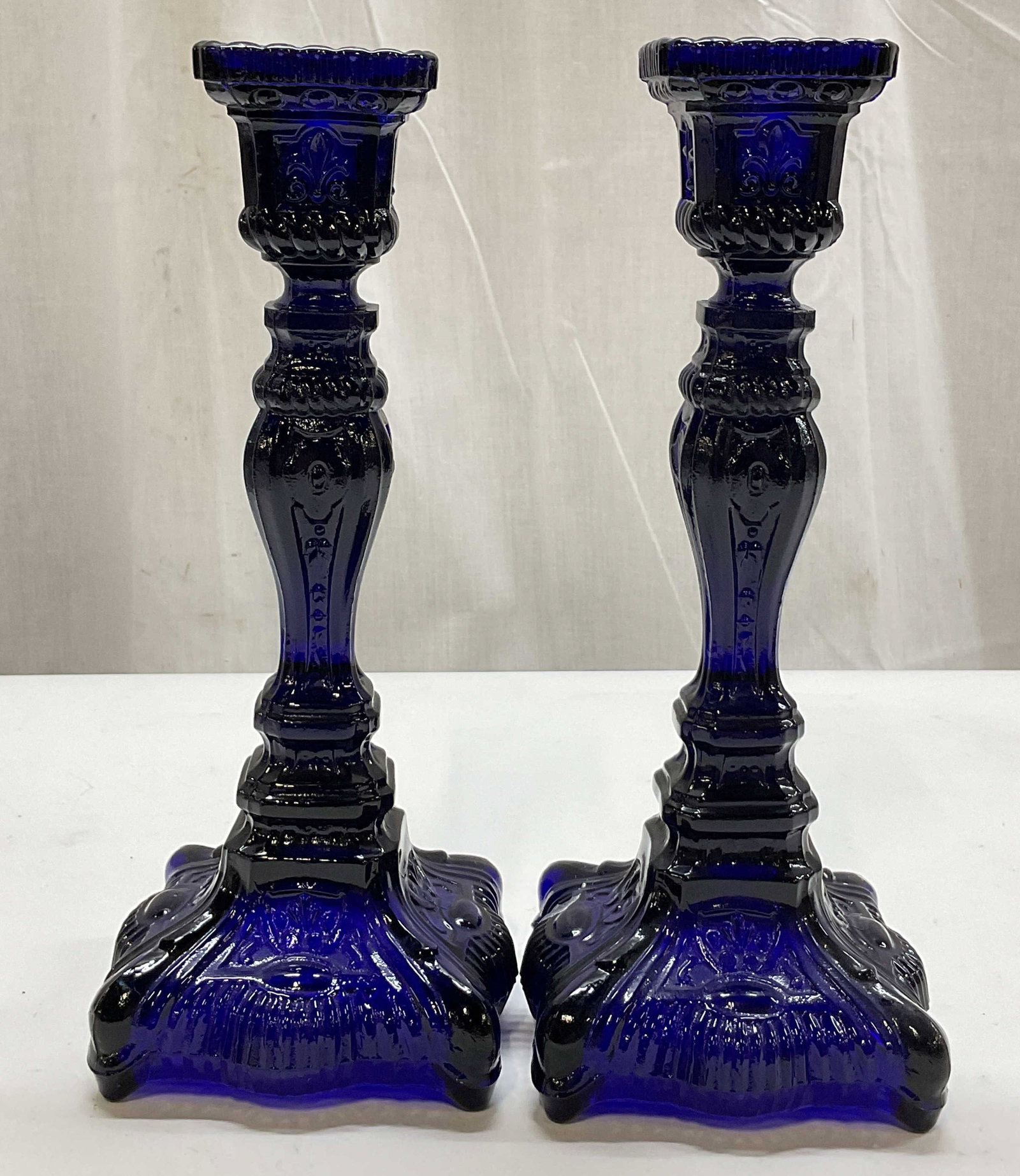 Pair Vintage Cobalt Blue Glass Candlesticks: Pair of vintage cobalt blue glass candlesticks. Square base with scalloped edge. Faceted and ribbed surface. Embossed scroll and floral motifs. Square top. Ornate design. Measures approx 4 x 4 x 10 in