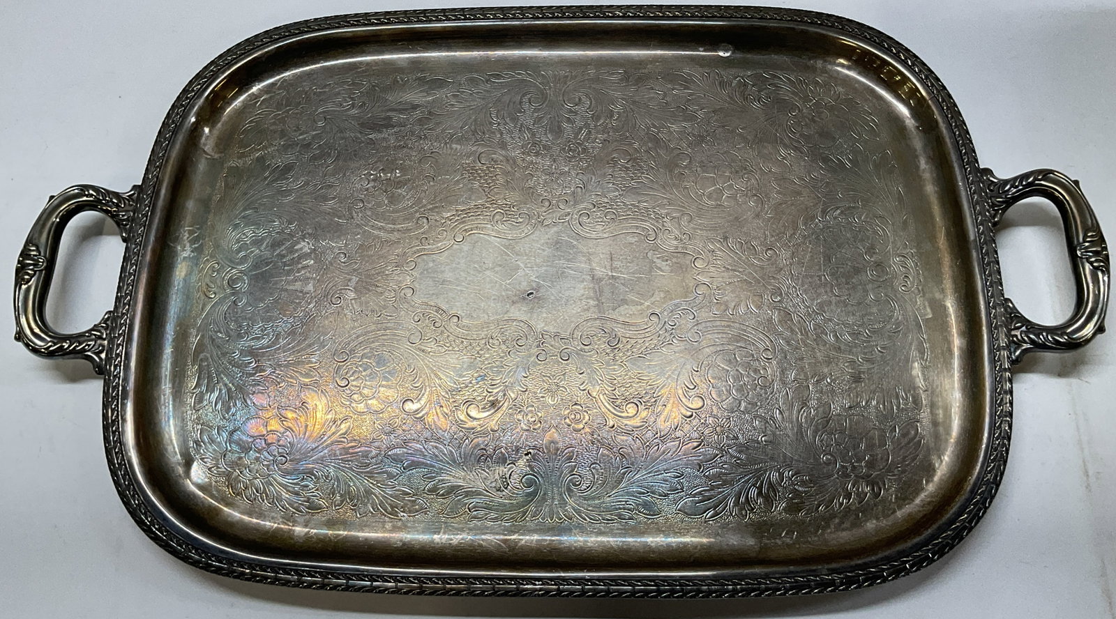 Vintage Silver Plated Footed Serving Tray 26in: Vintage silver plated footed serving tray. No visible markings. Scroll designed handles on either side. Round shape. Decorative rope edge rim. Embossed floral and scroll motifs. Feet on underside. Mea