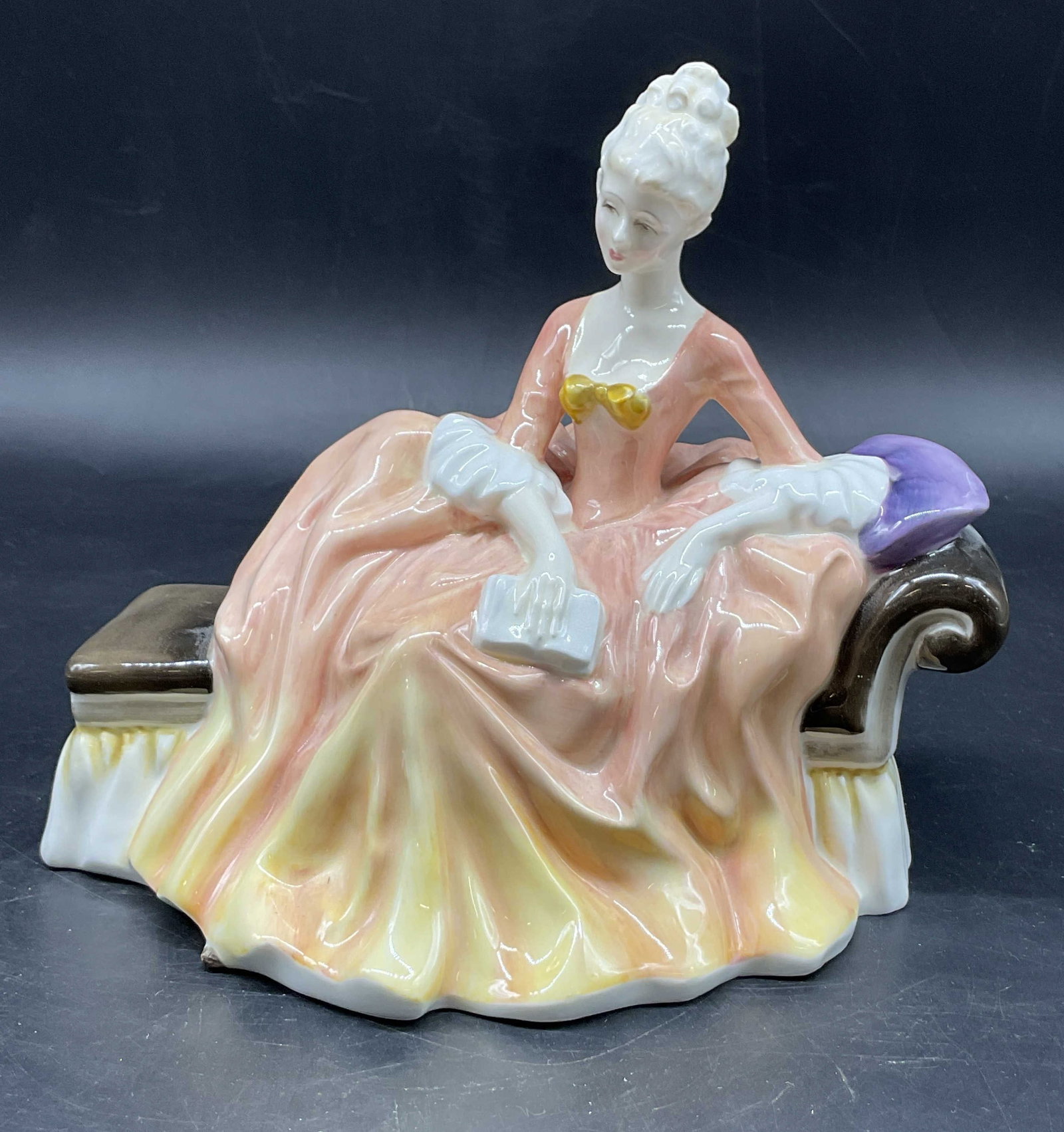 Royal Doulton Bone China Reverie Figure England: Royal Doulton bone china Reverie figure. Marked on underside, made in England. Figure is titled Reverie. Features a woman figure in a peach colored gown holding a book while reclining on a chaise loun